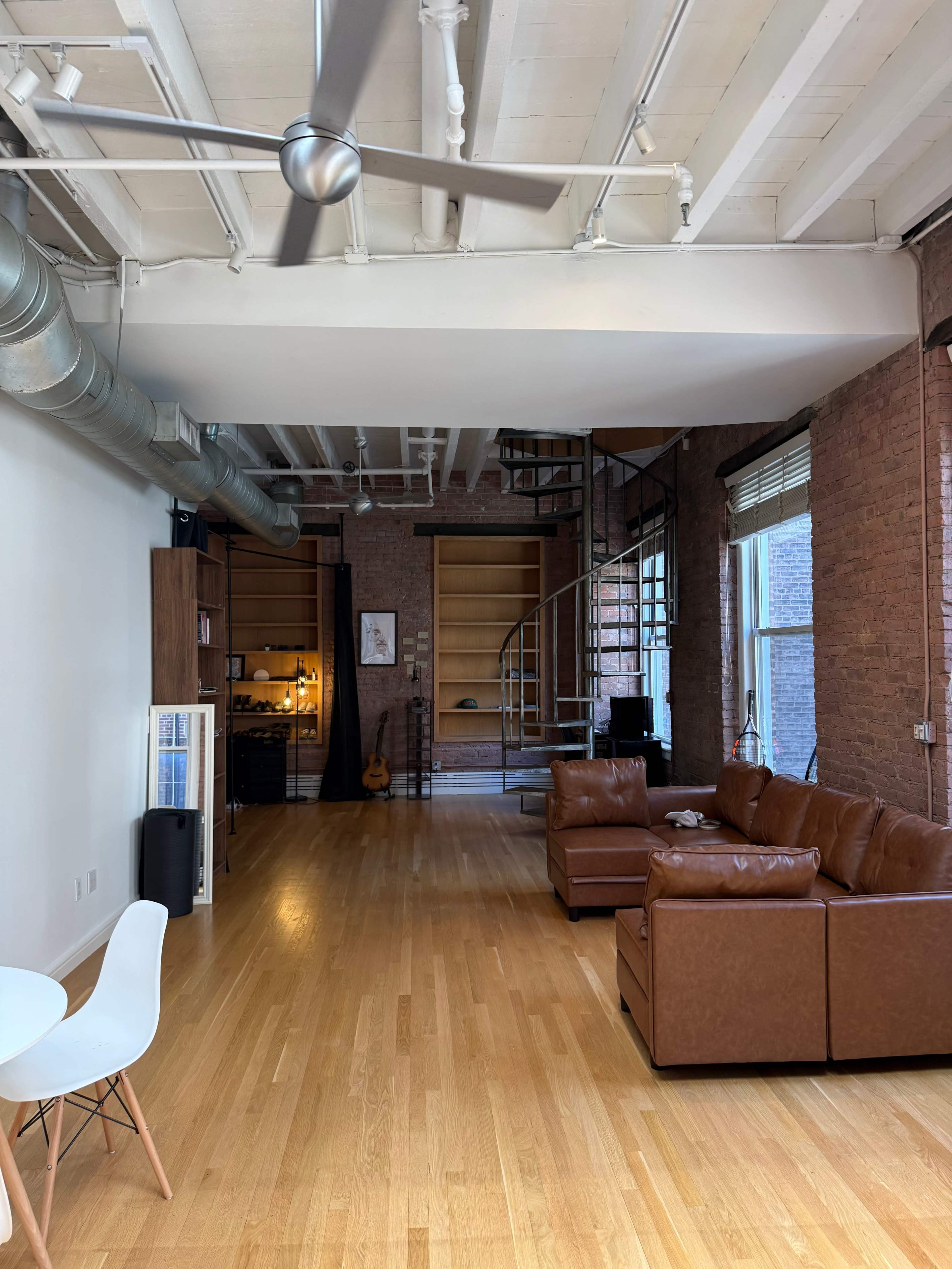The image shows a spacious living area with wooden flooring, a brown leather sectional sofa, and a spiral staircase leading to a loft space, set against exposed brick walls.