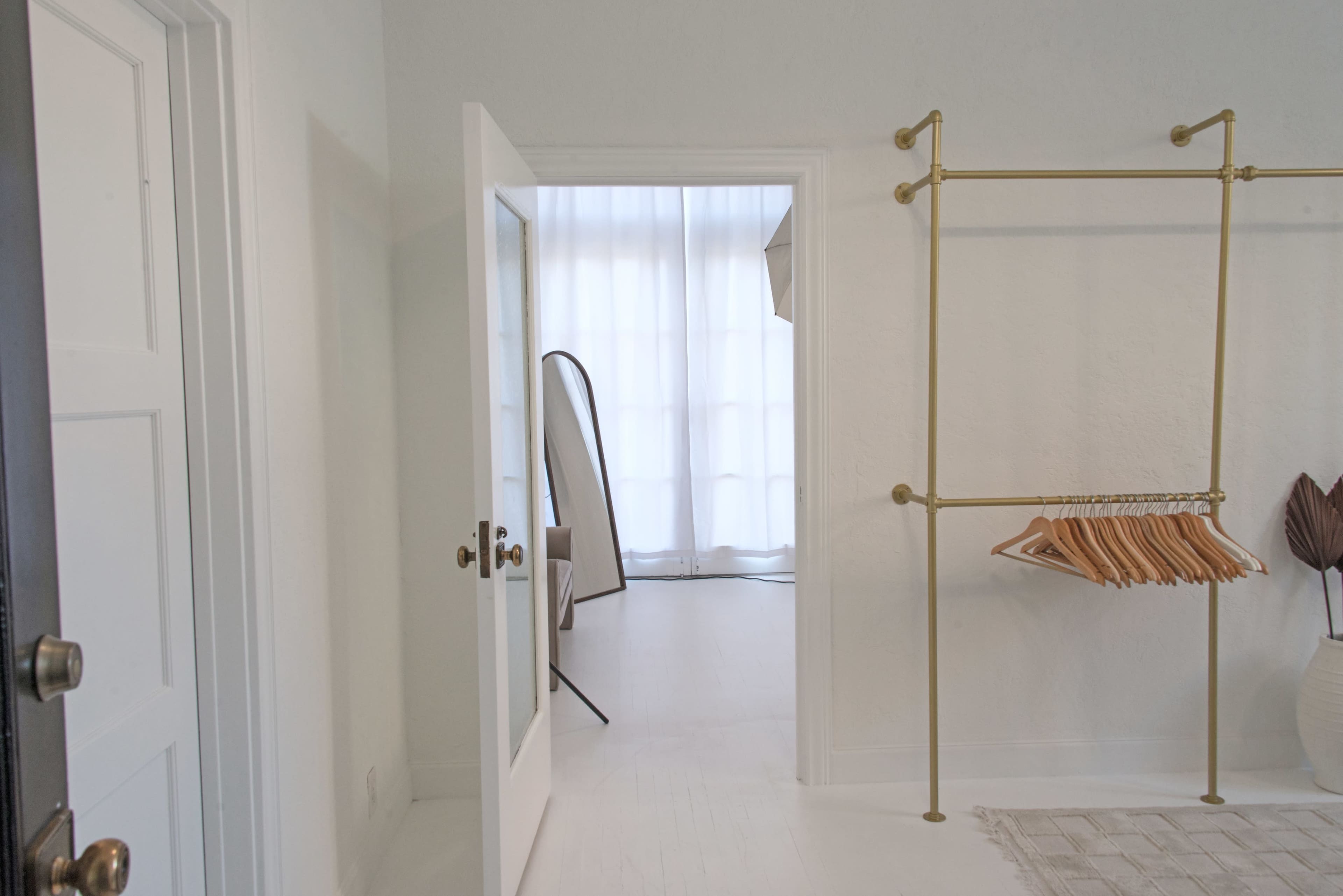 A white-walled room with an open door leads to another area featuring soft natural light, a clothing rack with hangers, and simple decor.