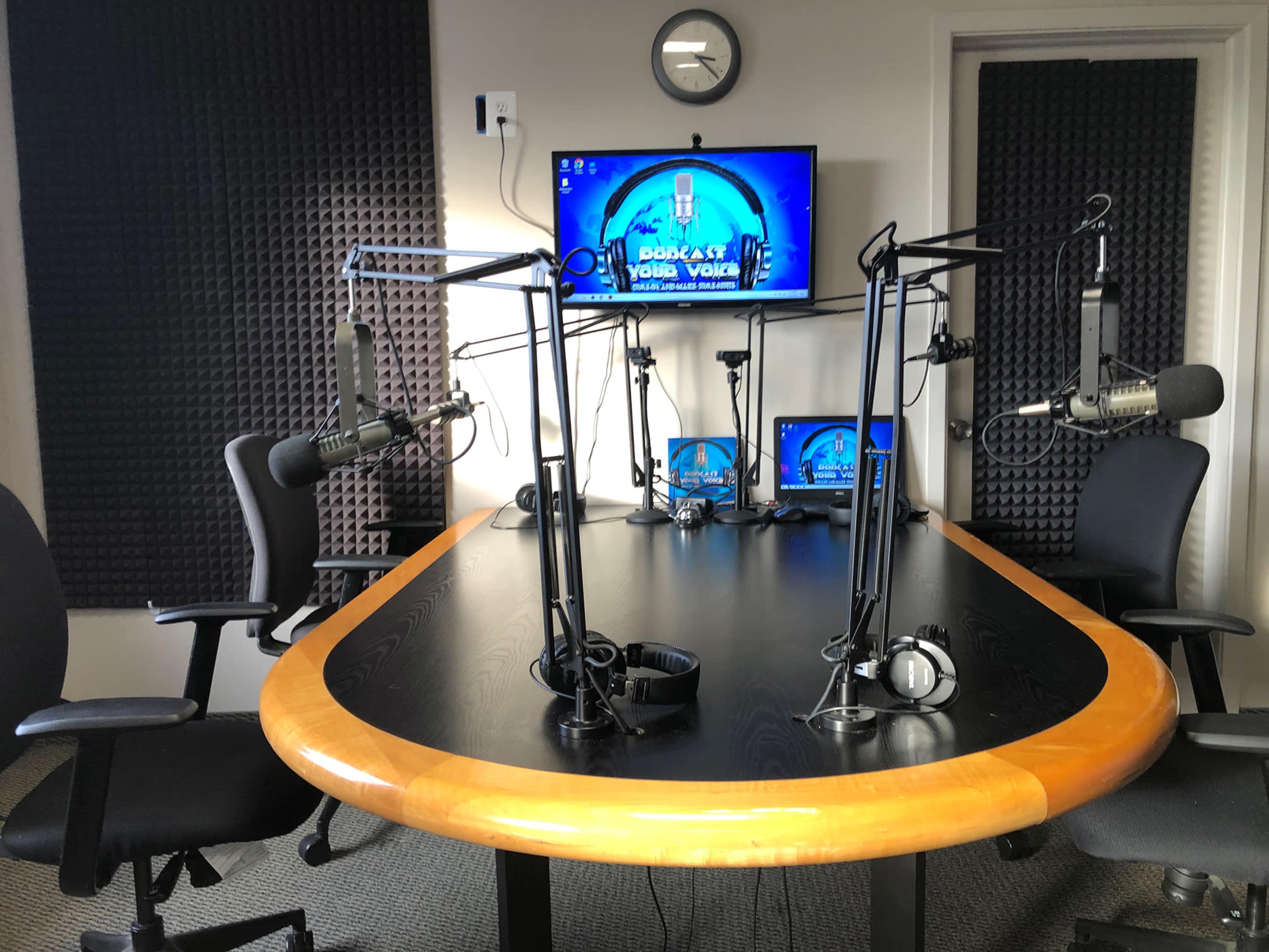The image shows a podcast studio featuring a large black table with microphones on adjustable arms, two monitors displaying the podcast logo, and soundproofed walls.