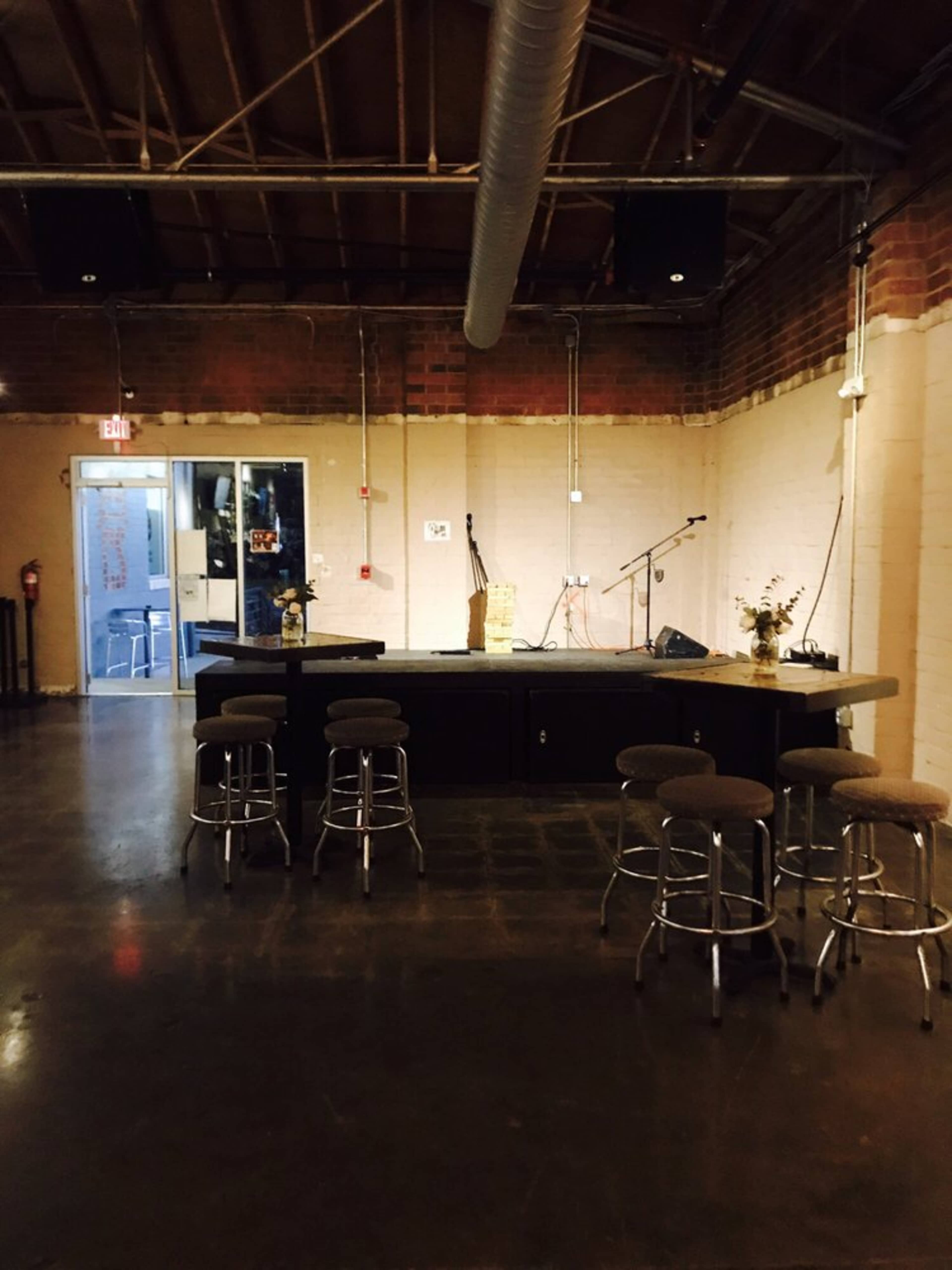 The image shows a sparse performance space with a small stage, an acoustic guitar stand, and several barstools aligned in front of the stage area.