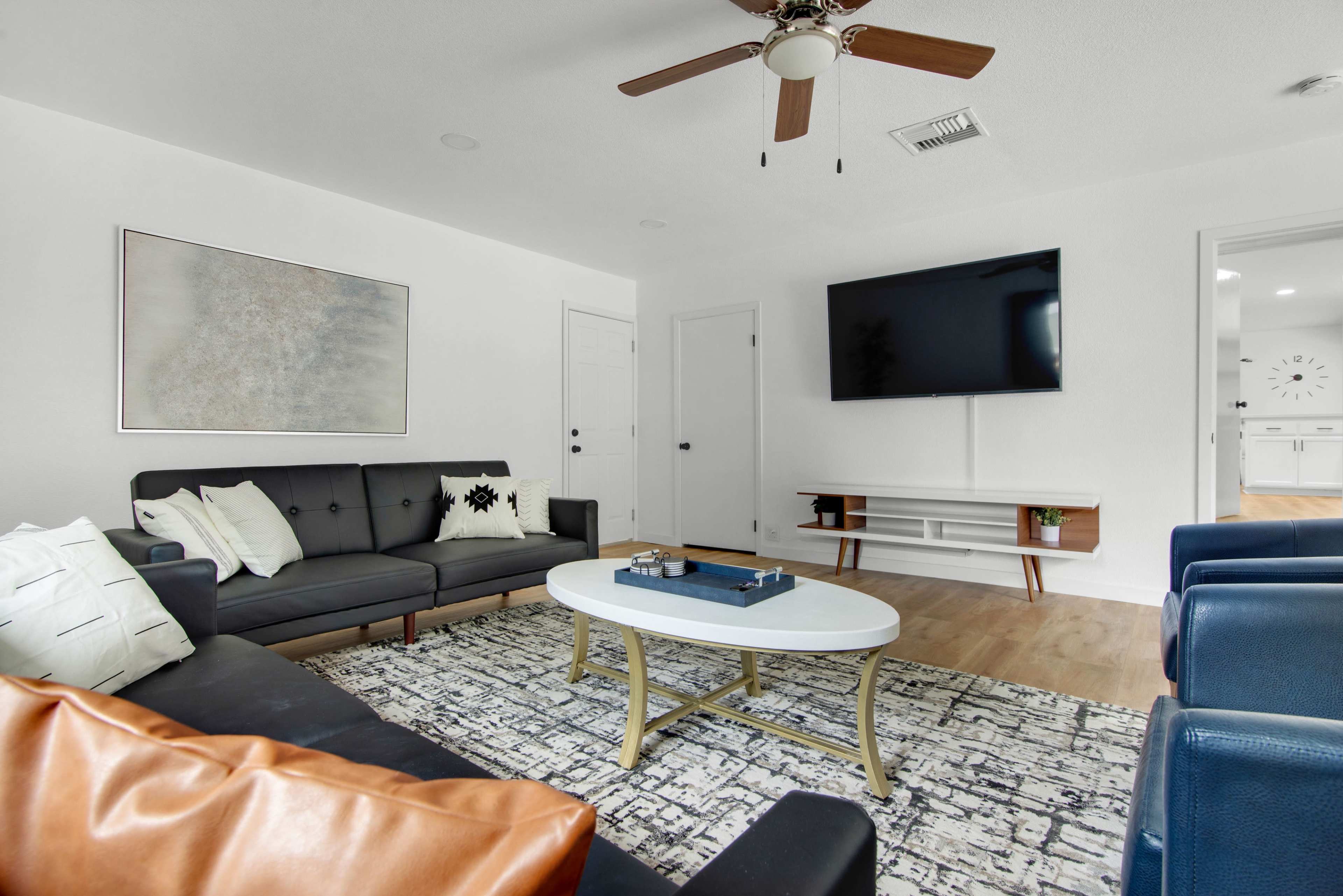 A modern living room features a dark sectional sofa, a central coffee table, and a wall-mounted television, with wooden flooring and a light color scheme.