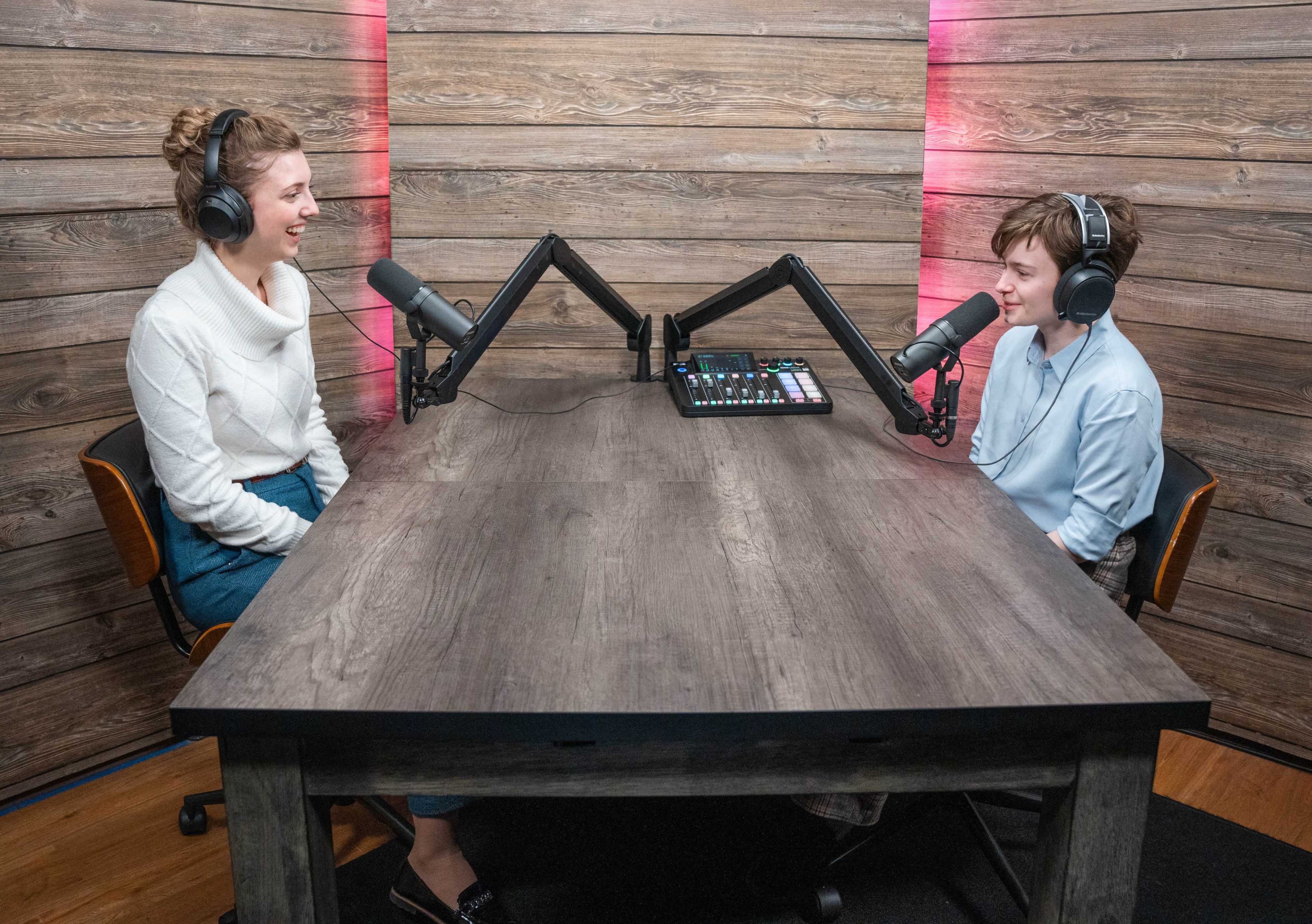 Two individuals sit at a wooden table equipped with microphones and audio equipment, engaged in conversation in a recording studio.