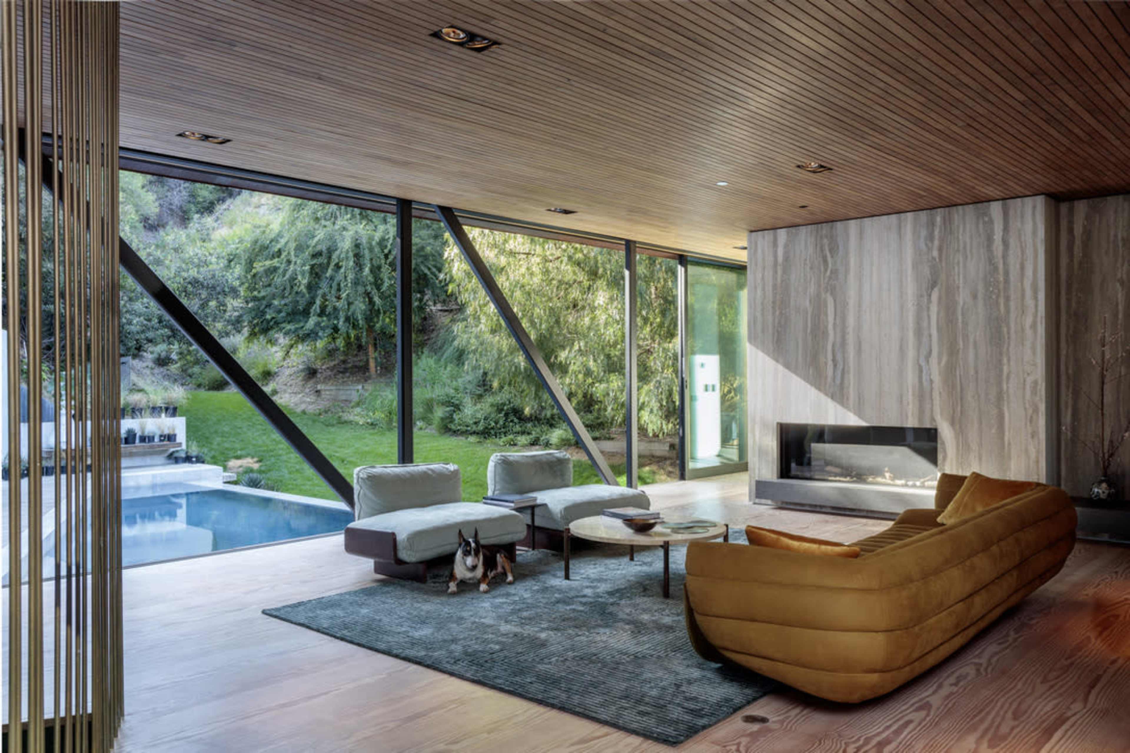 The image shows a modern living room with a large window overlooking a green outdoor area, featuring a yellow couch, a coffee table, and a fireplace on one wall.