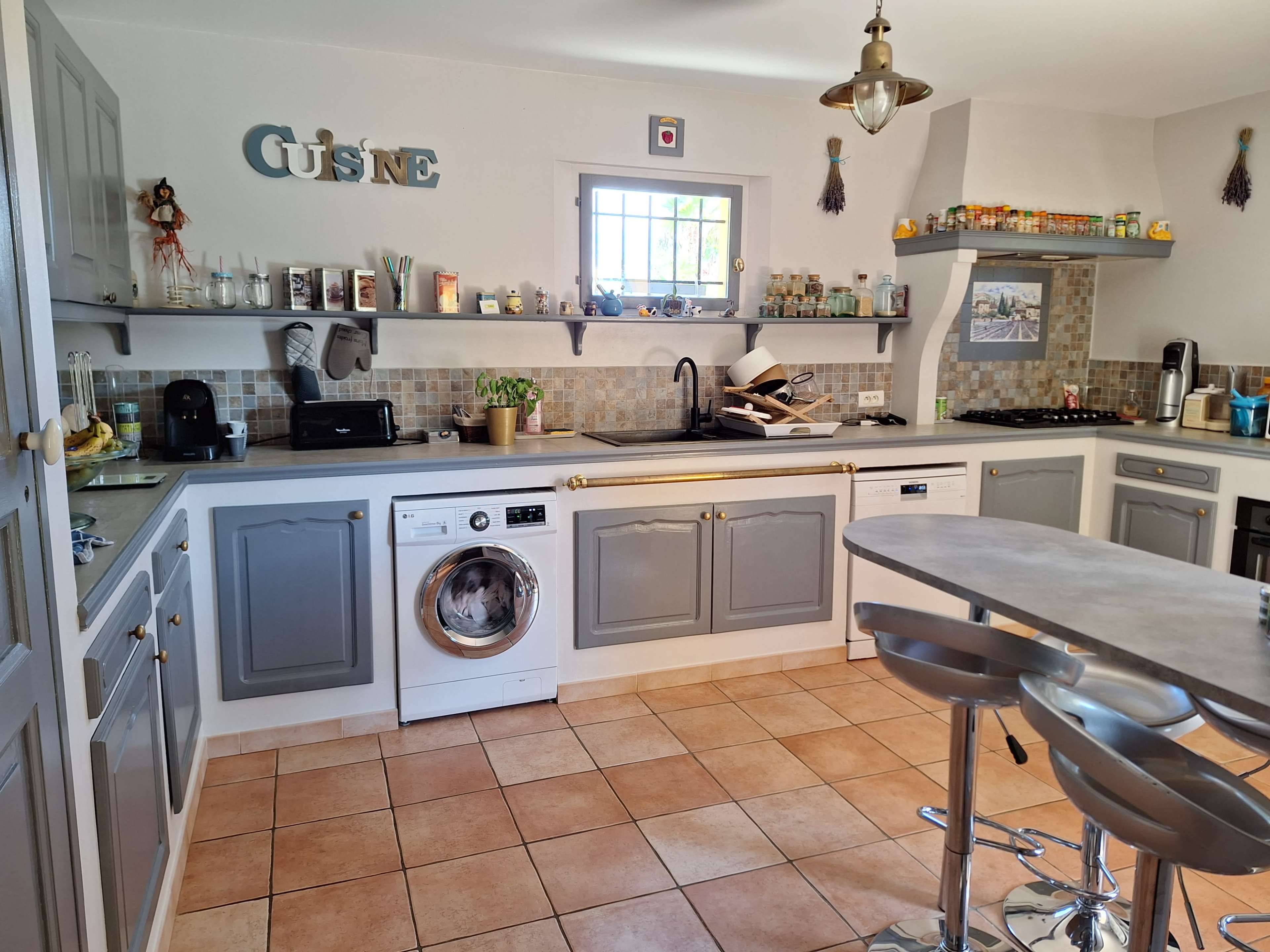 The image shows a modern kitchen with gray cabinets, a washing machine, and a tiled floor, featuring a variety of kitchen appliances and decorative items on shelves.