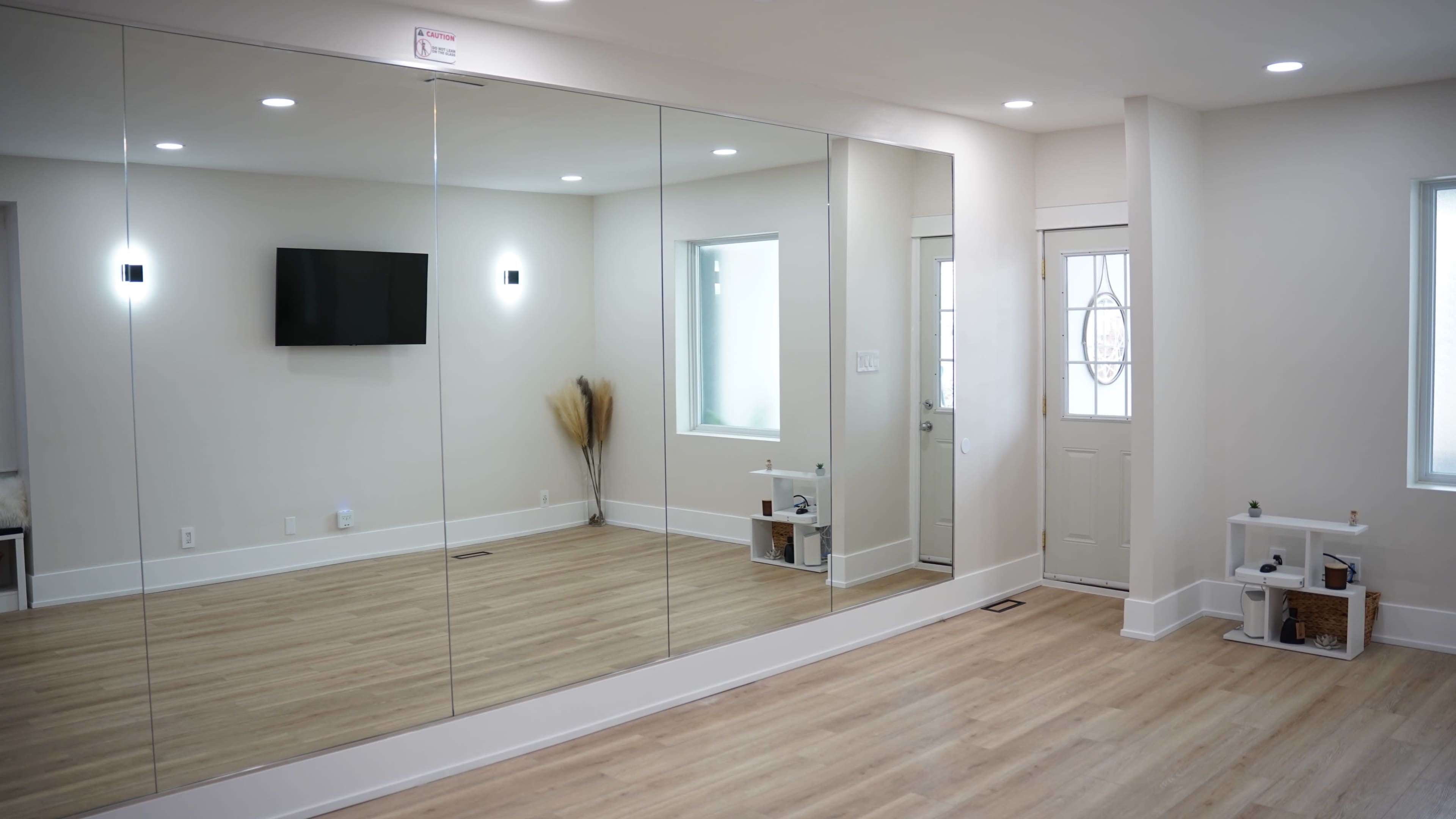 A spacious room features a large mirrored wall, a small television mounted on the opposite wall, and a front door with a window beside it.