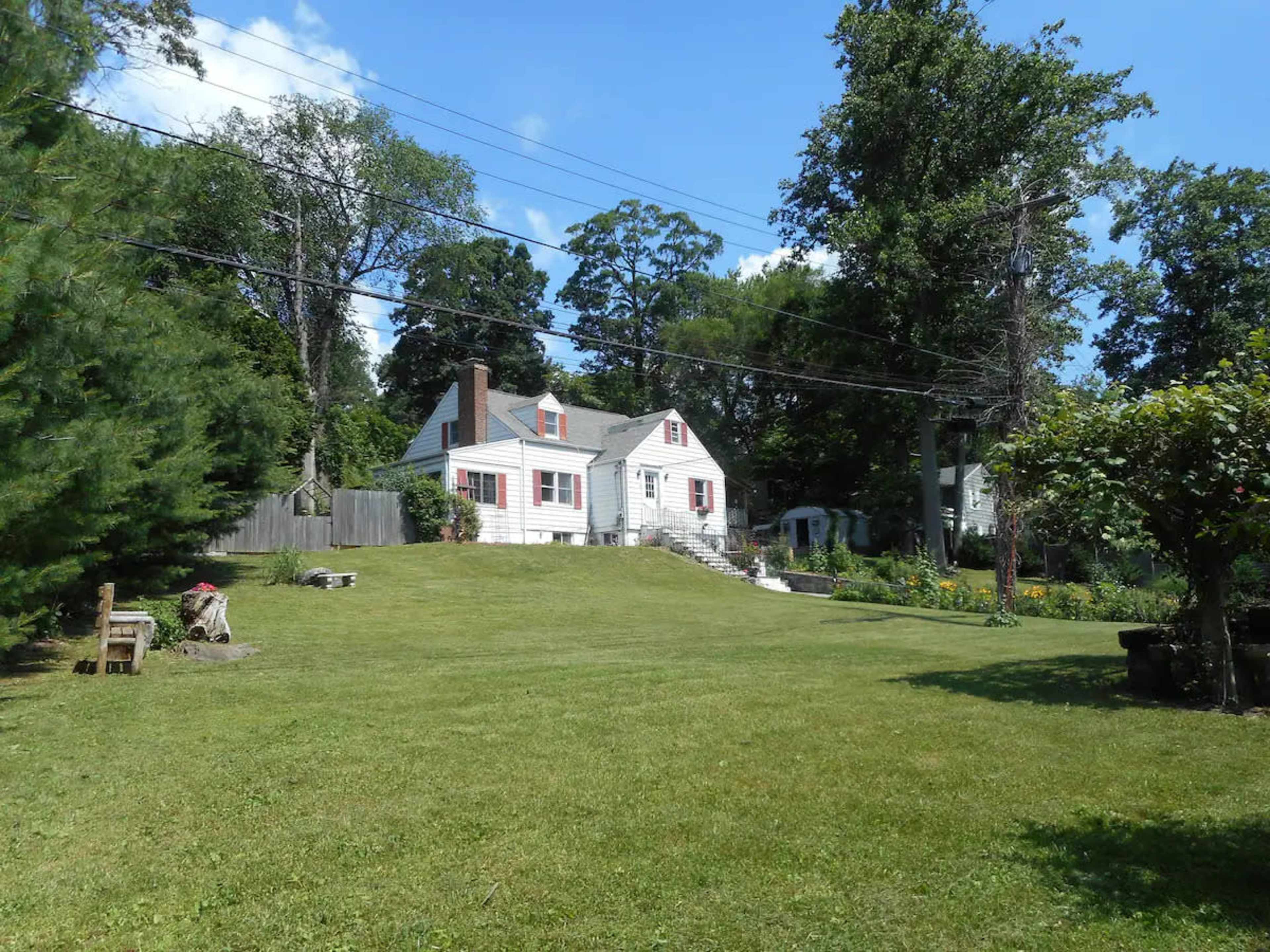 A single-story white house with a sloped roof is situated on a large grassy lawn surrounded by trees and electrical poles.