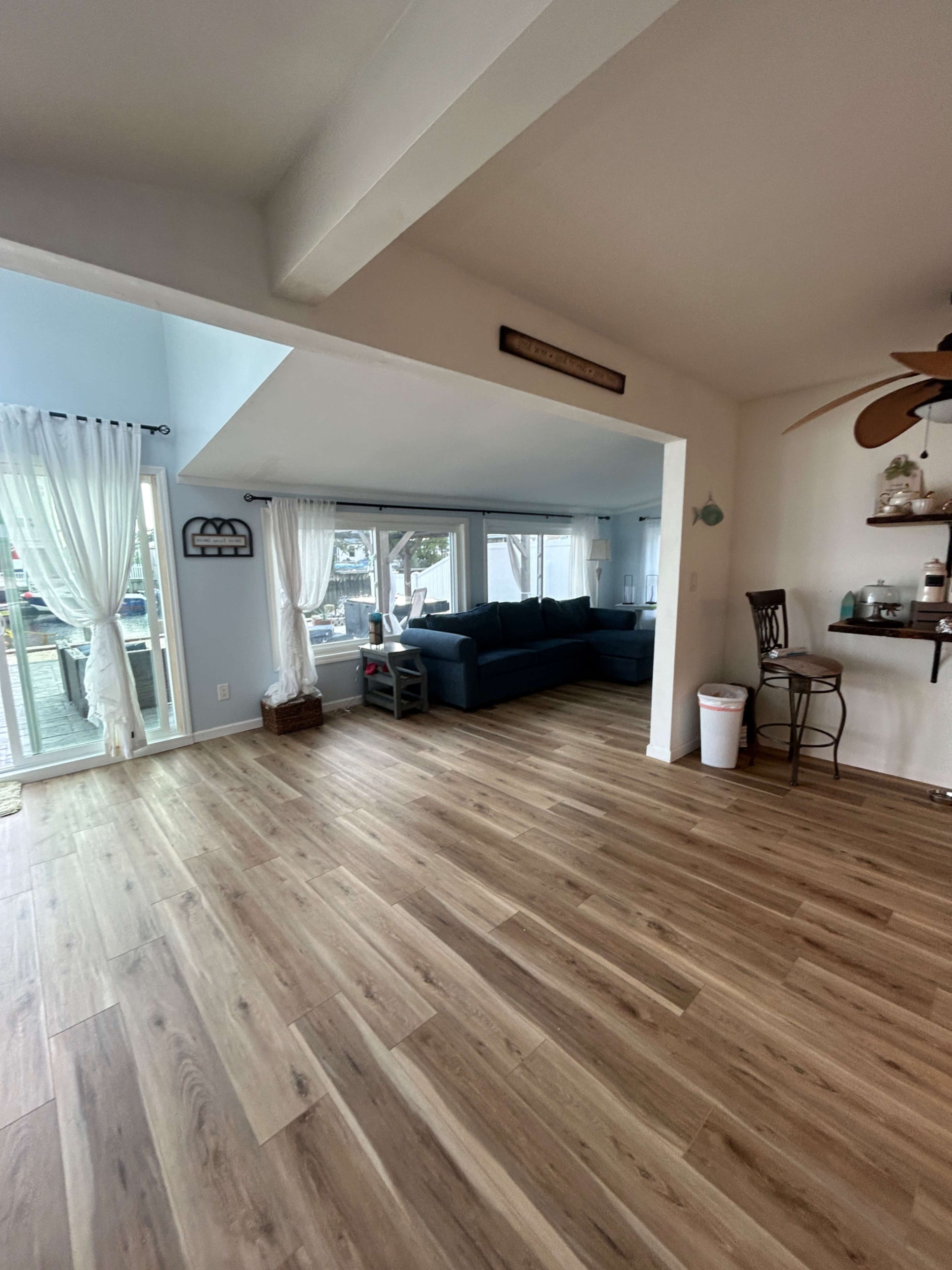 A spacious room features a large sectional sofa, large windows with sheer curtains, and hardwood flooring.