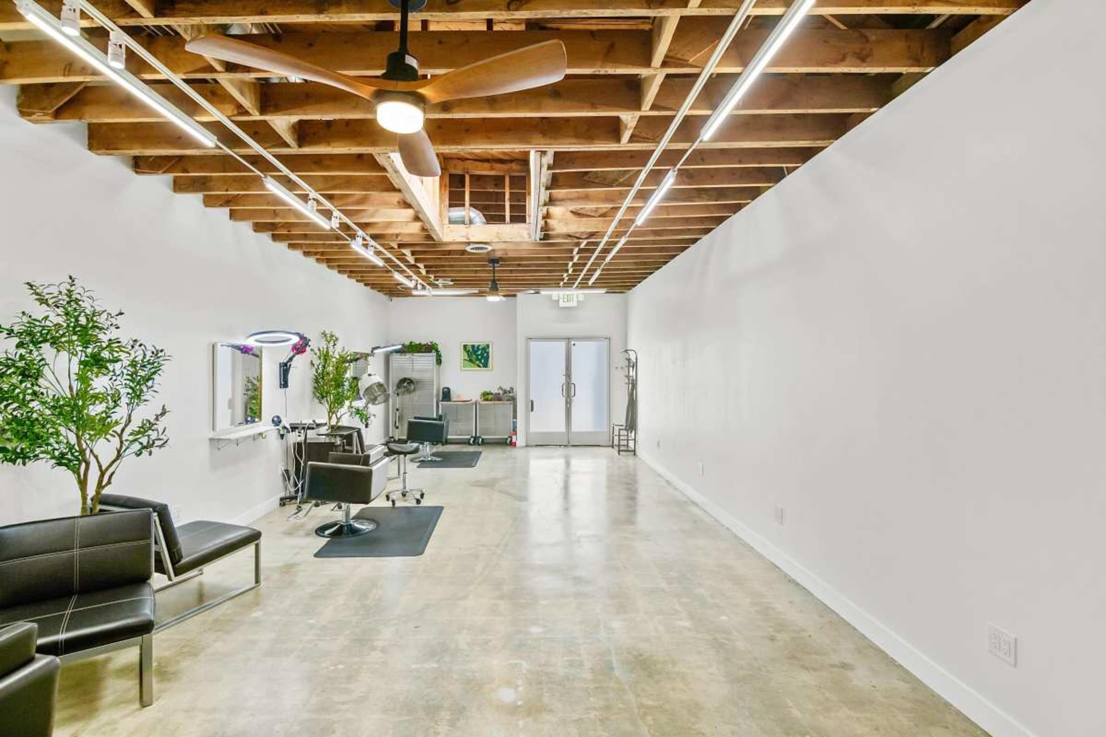 The image shows a spacious salon interior with a minimalistic design, featuring a black leather couch, salon chairs, mirrors, and a ceiling with exposed beams and modern lighting.