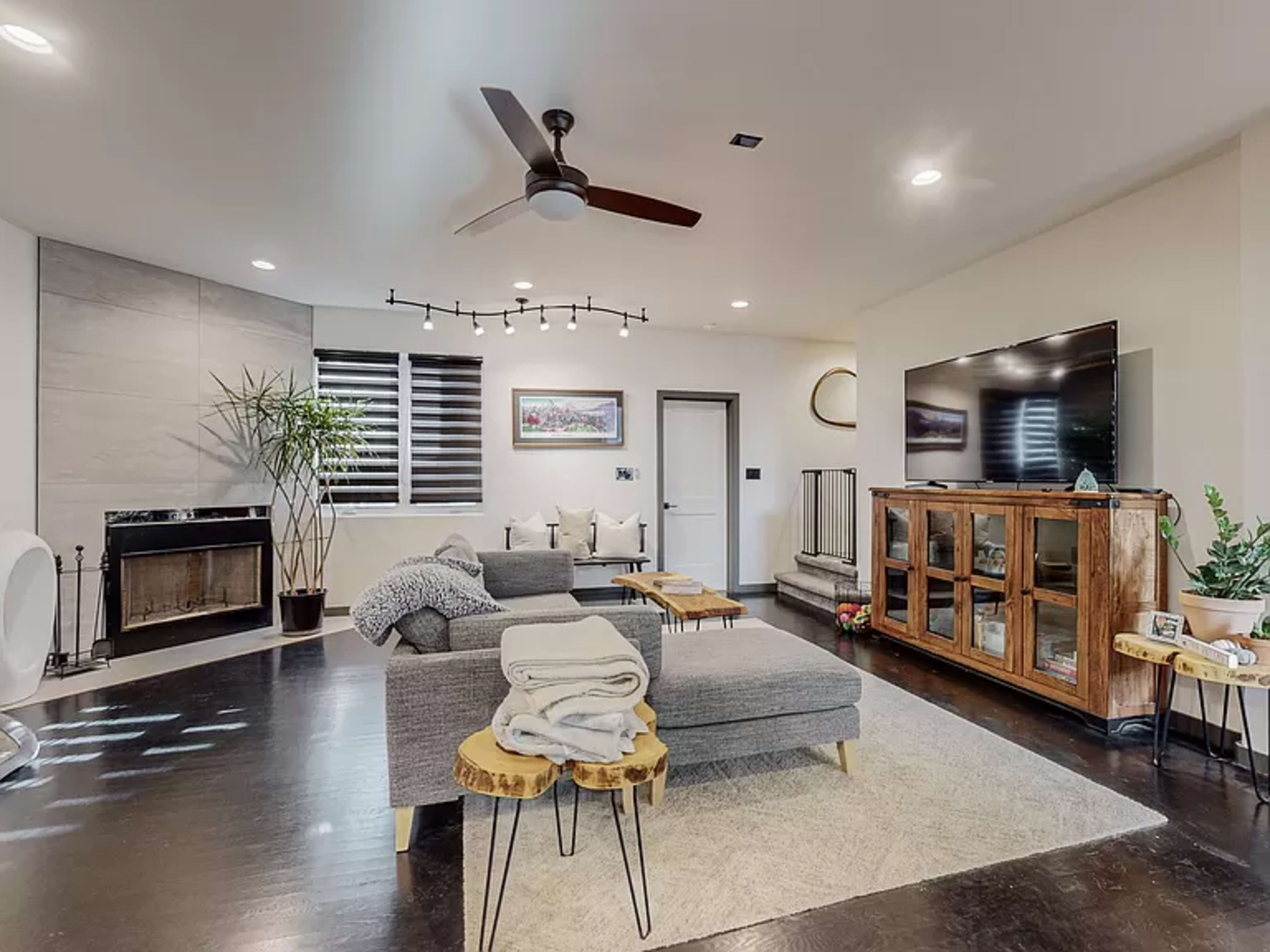 A modern living room featuring a comfortable gray sectional sofa, a large television mounted on the wall, and a wooden entertainment center, all set against a backdrop of hardwood flooring and natural light from the windows.