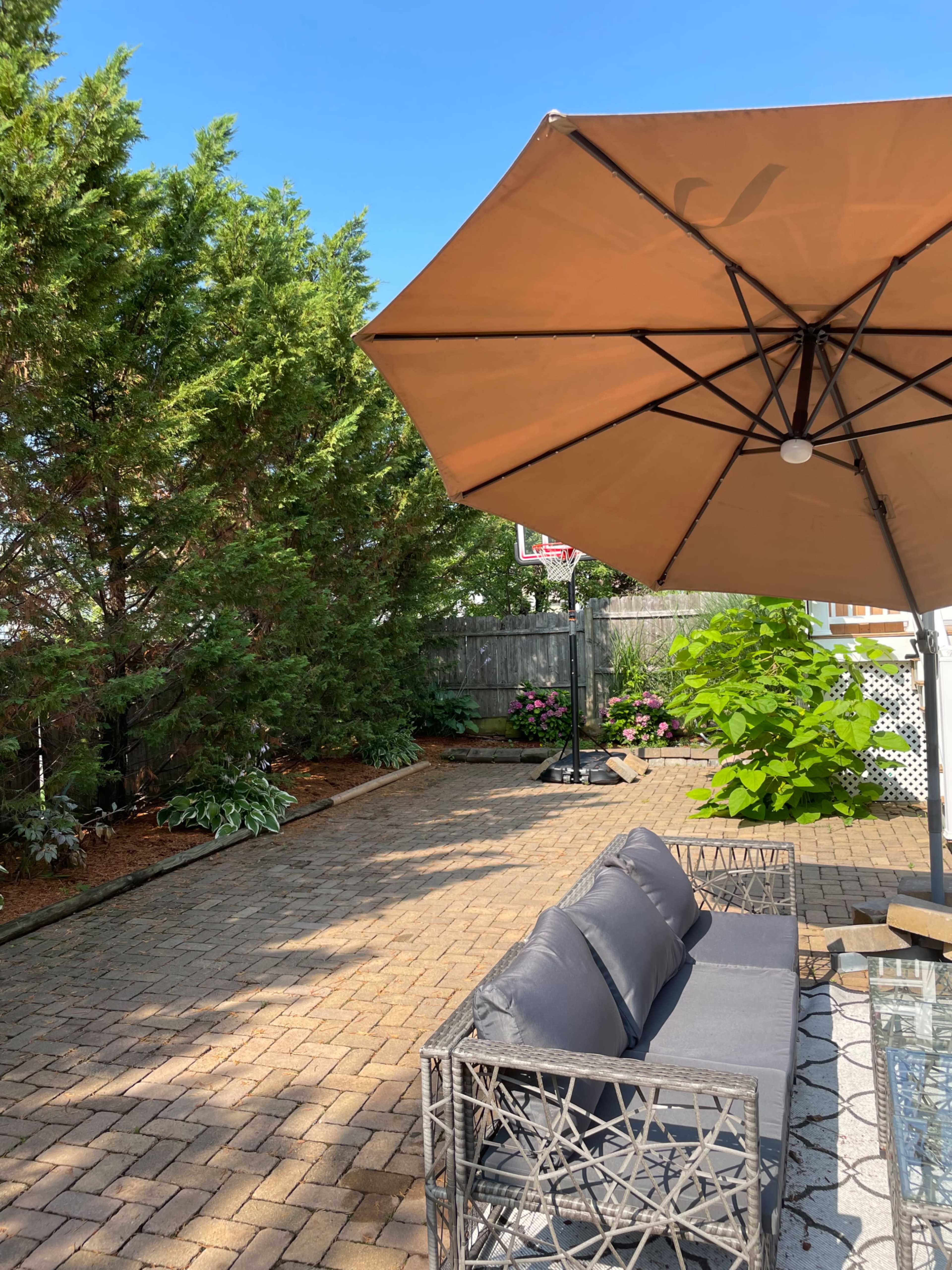 A patio area features a shaded seating arrangement under a large umbrella, surrounded by greenery and flowerbeds.