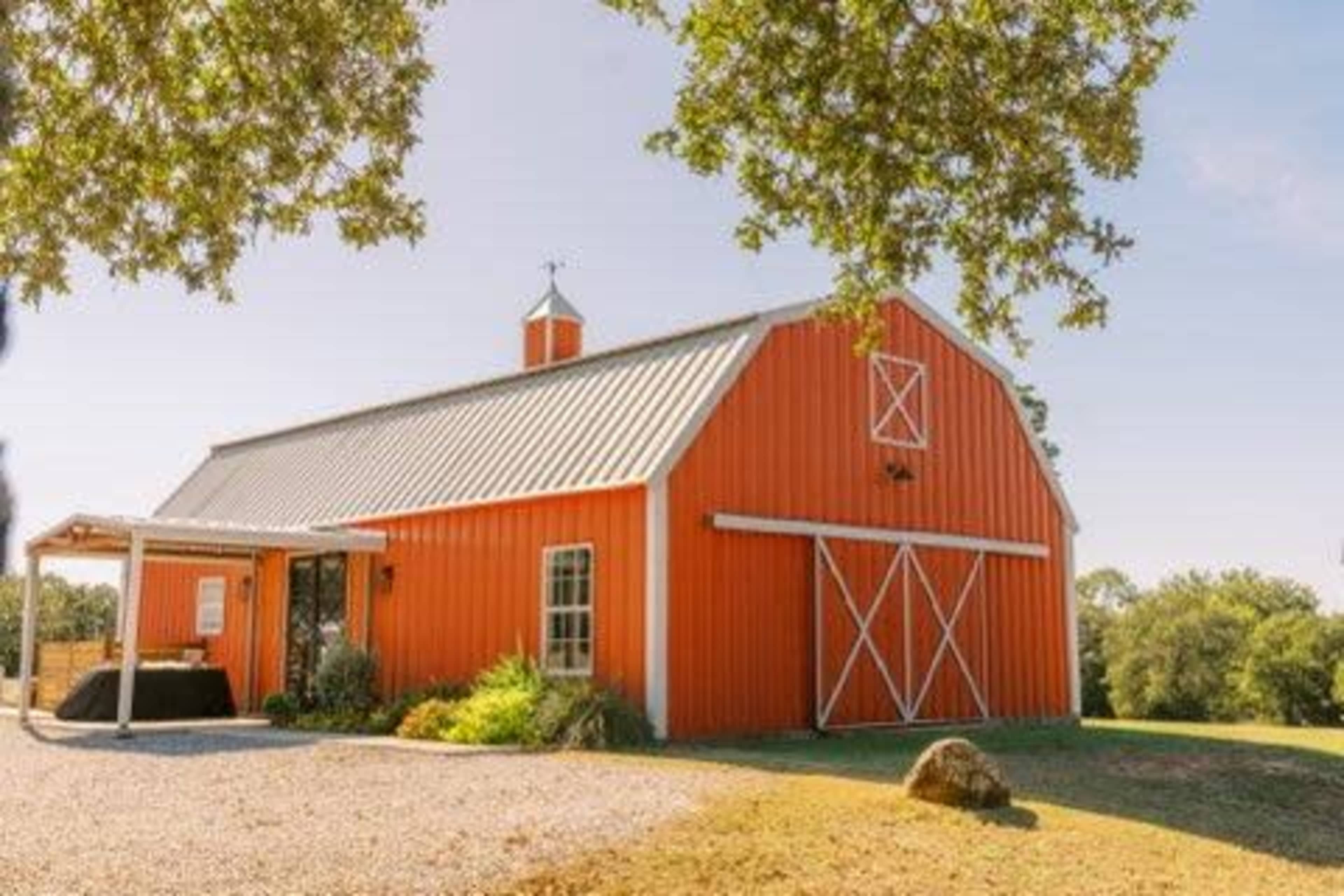 The image shows a bright orange barn with a metal roof located in a grassy area under a clear blue sky.