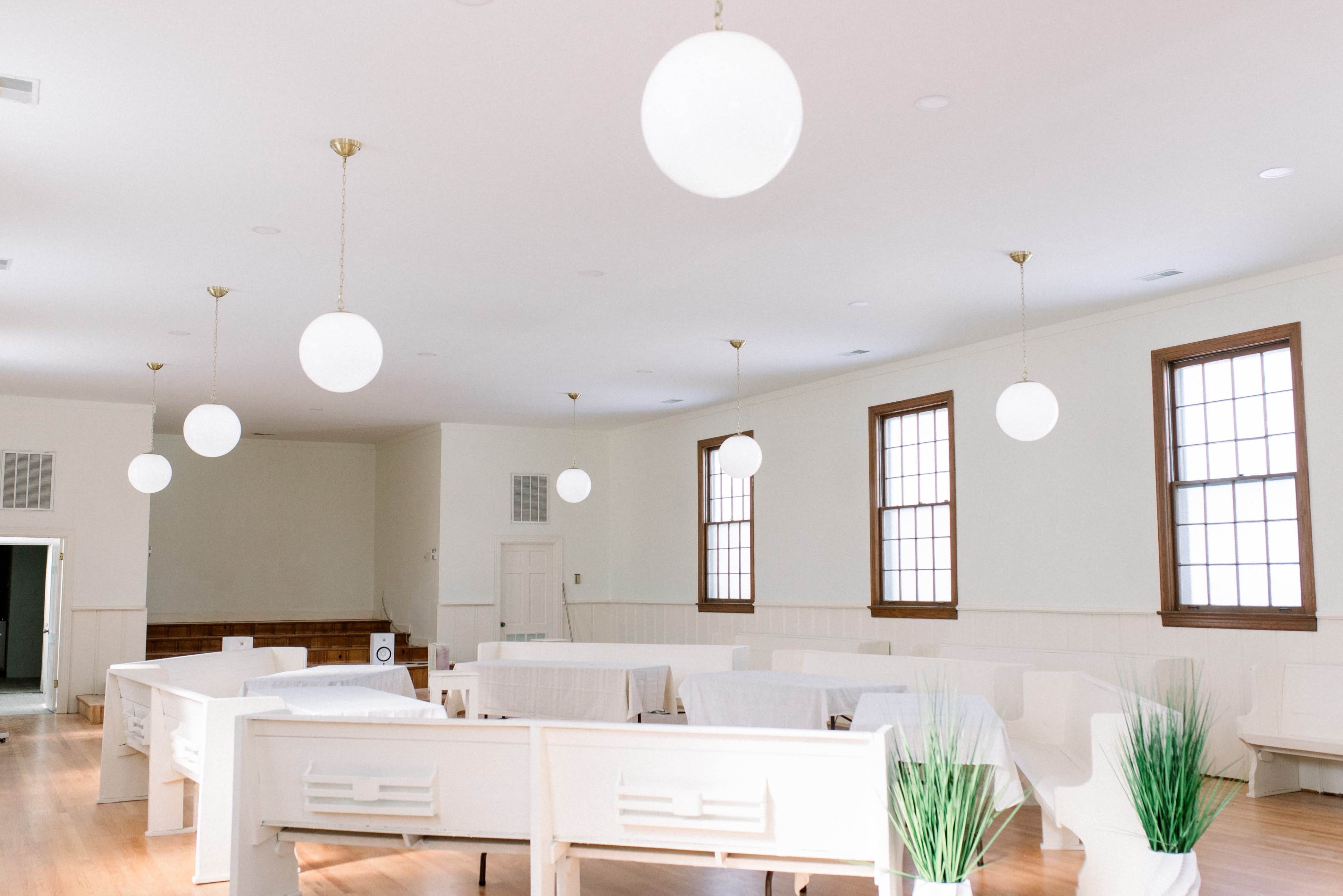 A minimalistic interior of a room with white walls, wooden floors, and round ceiling lamps, featuring several rows of white benches and green plants.
