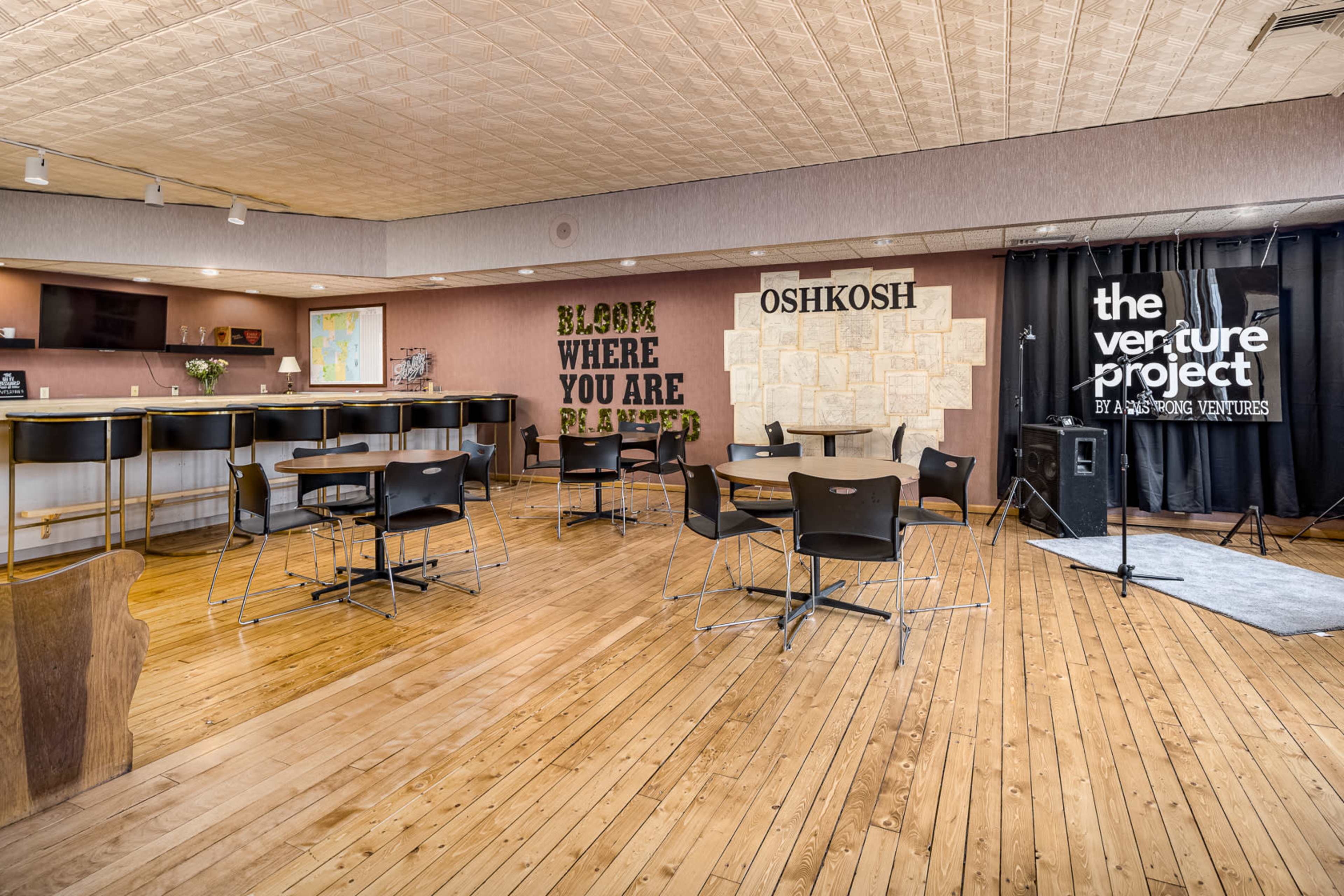 A spacious meeting room features wooden floors, several round tables with black chairs, and a backdrop with decorative text on the walls.