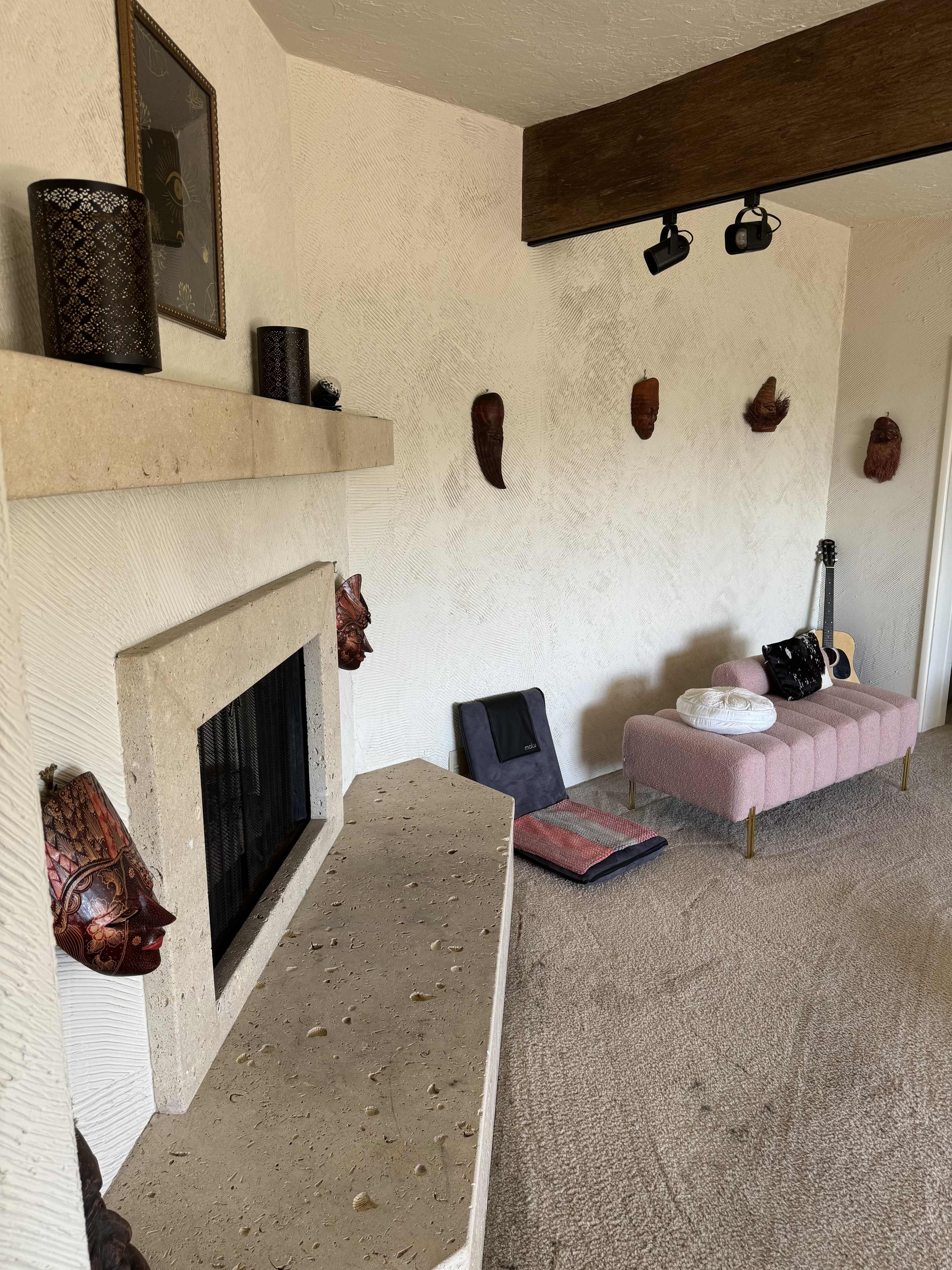A living room with a stone fireplace, a pink upholstered bench, and several decorative masks mounted on the walls.