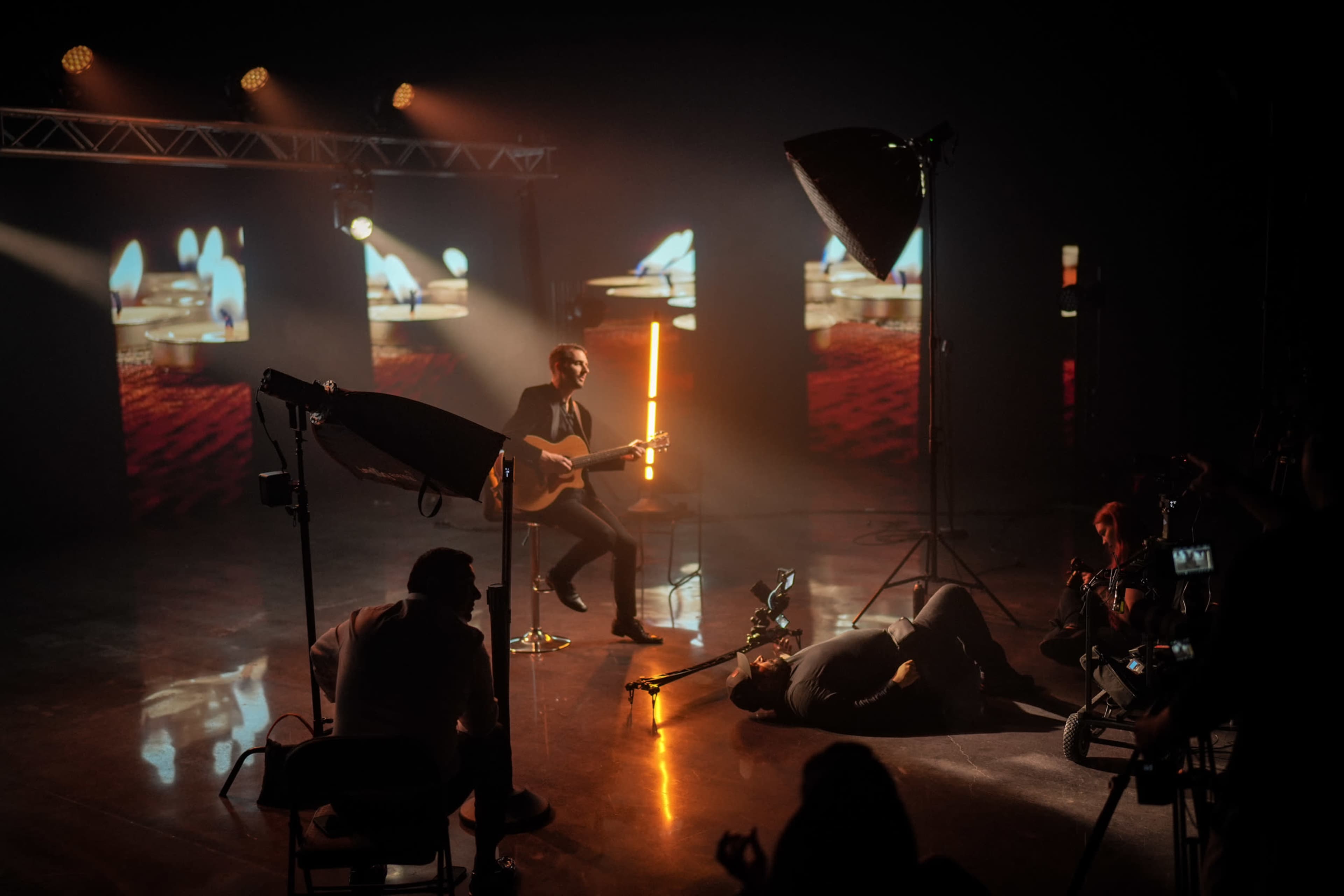 A musician plays guitar on stage with warm lighting while a camera crew captures the performance in a studio setting.