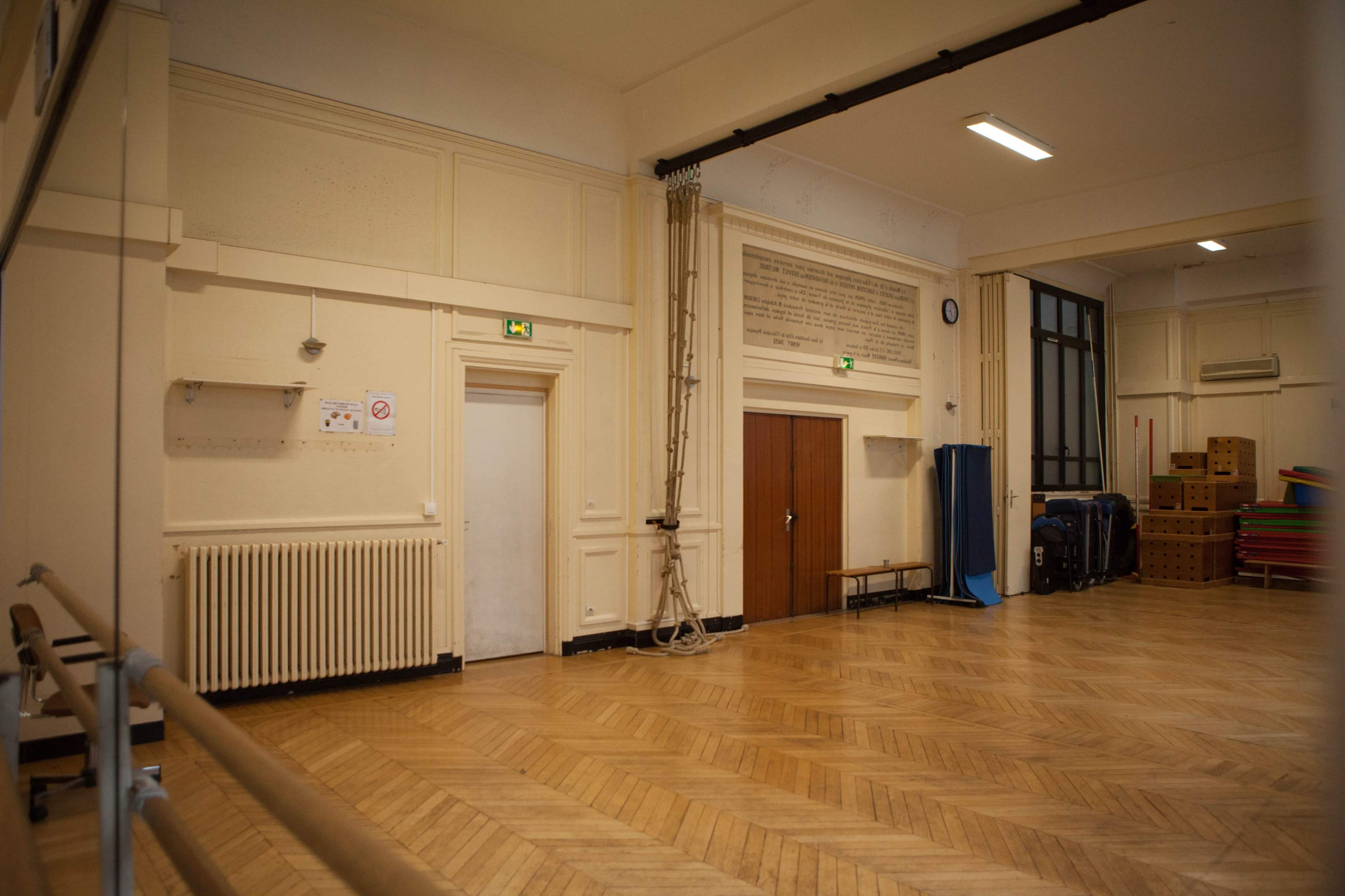 The image shows the interior of a spacious room with wooden flooring, a ballet barre along one wall, and various storage items, including stacked mats and boxes.