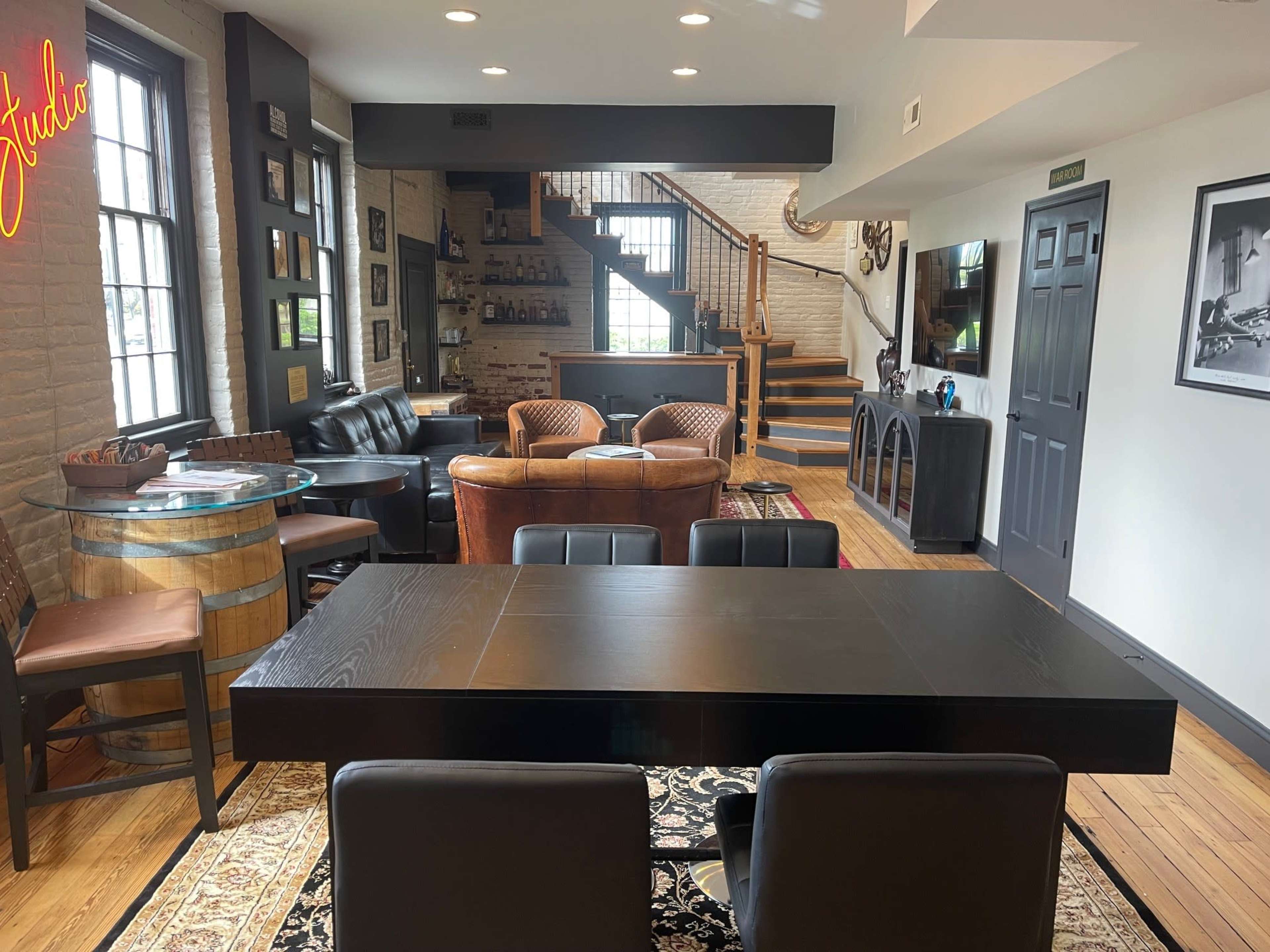 The image shows a contemporary living space with a dining table at the forefront, leather seating areas, a staircase in the background, and bar shelves along the wall.