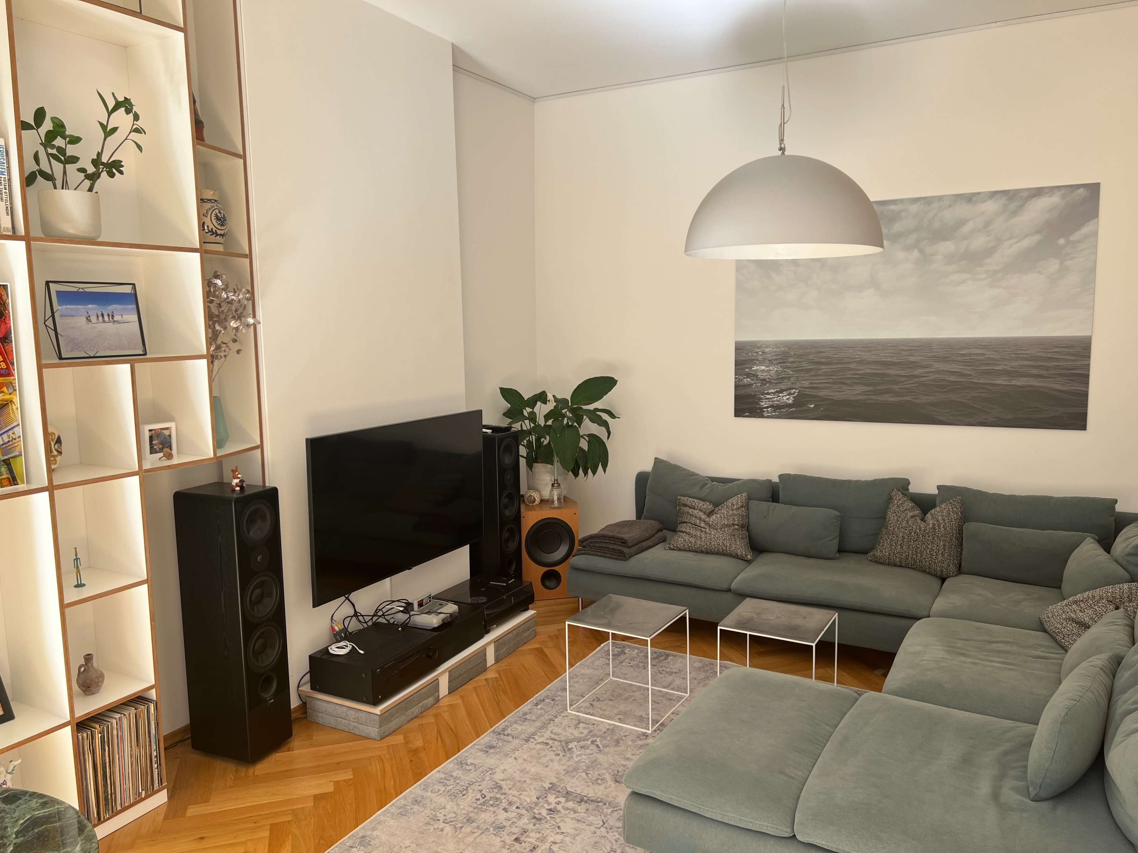 A spacious living room features a large sectional sofa, a wall-mounted television, and a decorative shelf with plants and books.