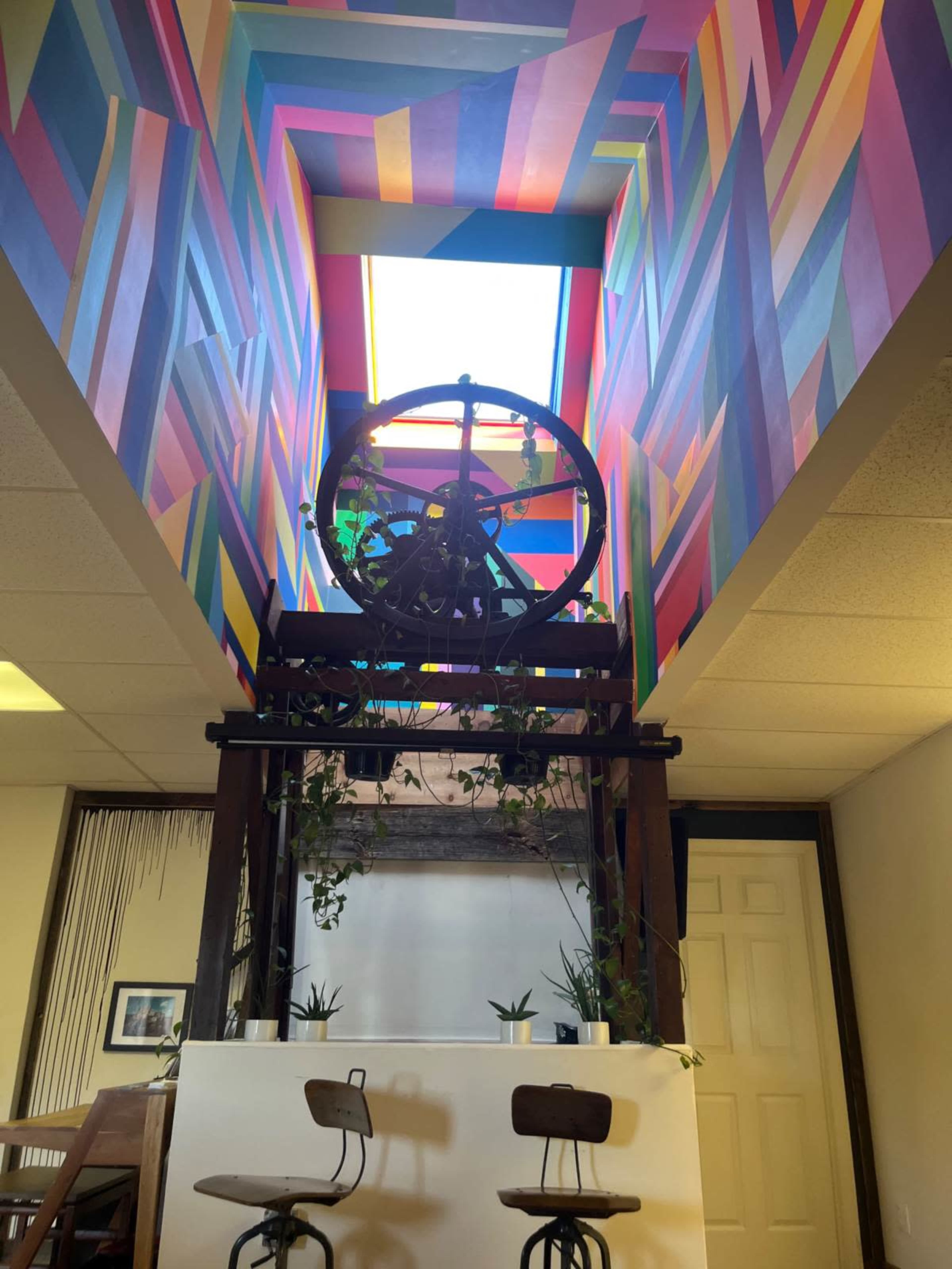 The image shows a brightly colored corridor with a geometric mural on the ceiling and a wooden structure with plants and two chairs below it.