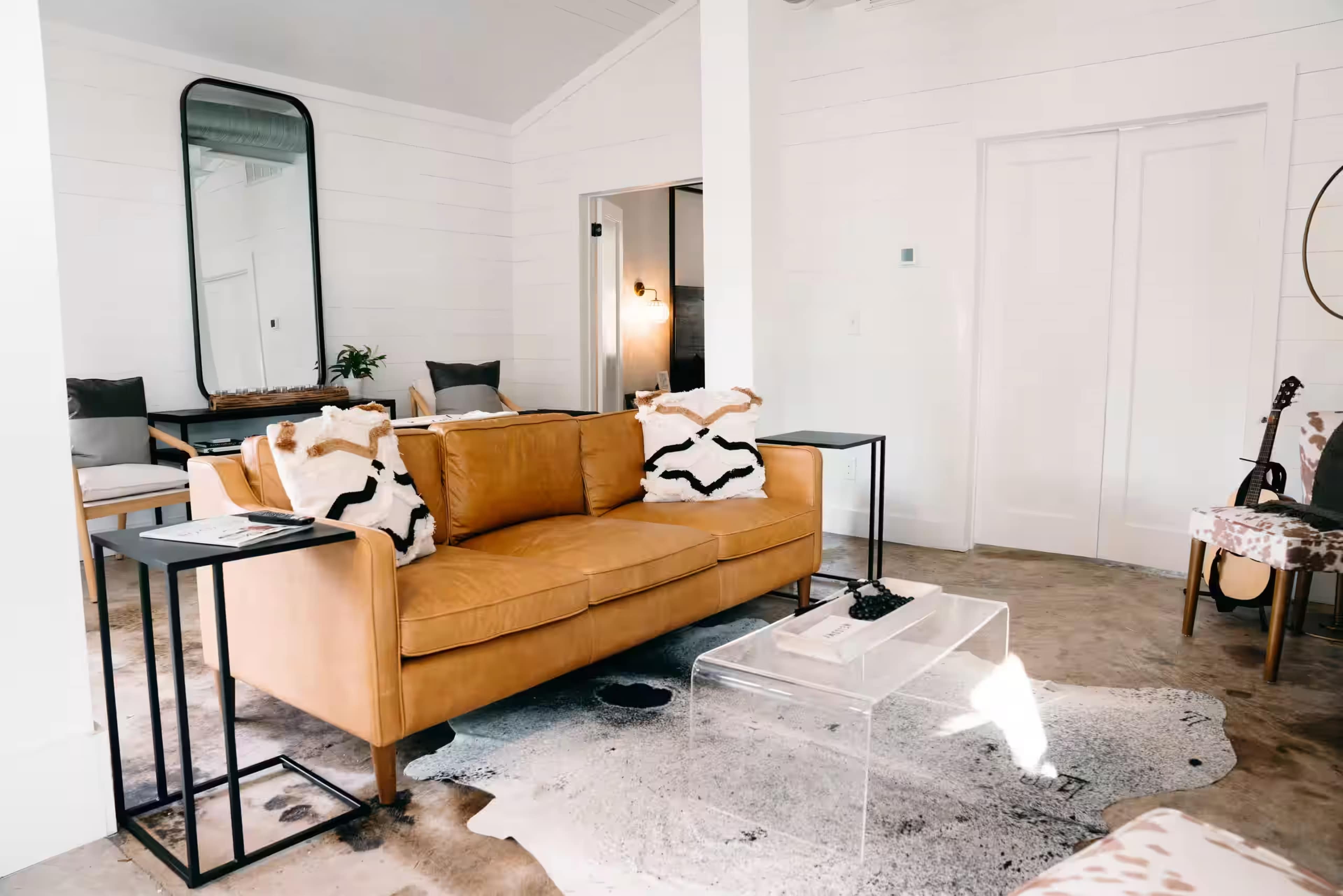 The image shows a modern living room with a brown leather sofa, transparent coffee table, and a decorative rug in a bright, minimalist space.