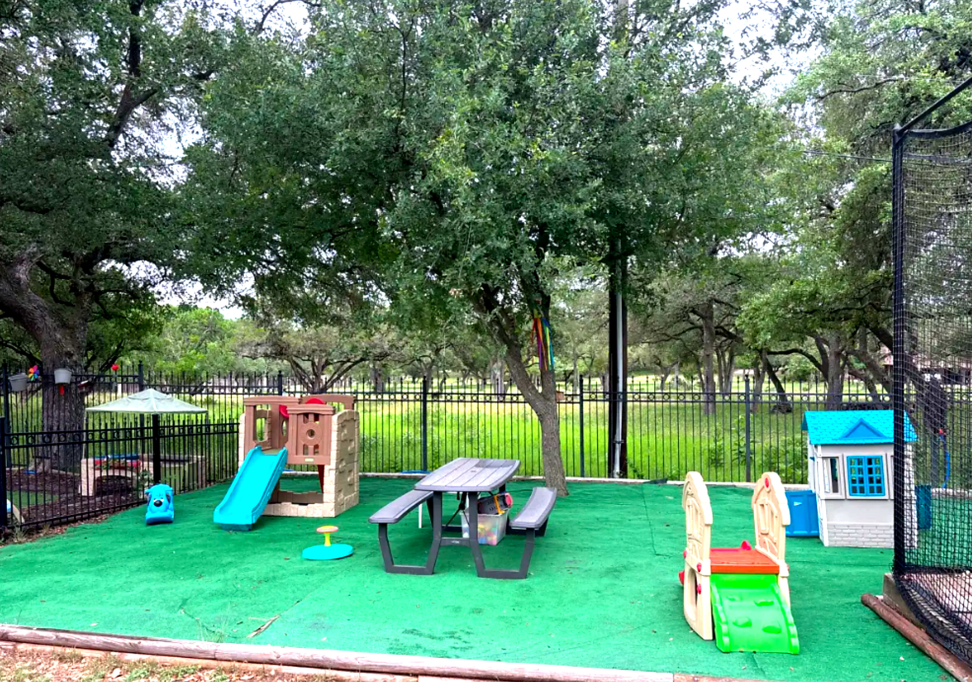 The image shows a playground area with a slide, a picnic table, and a small playhouse, surrounded by green grass and trees.