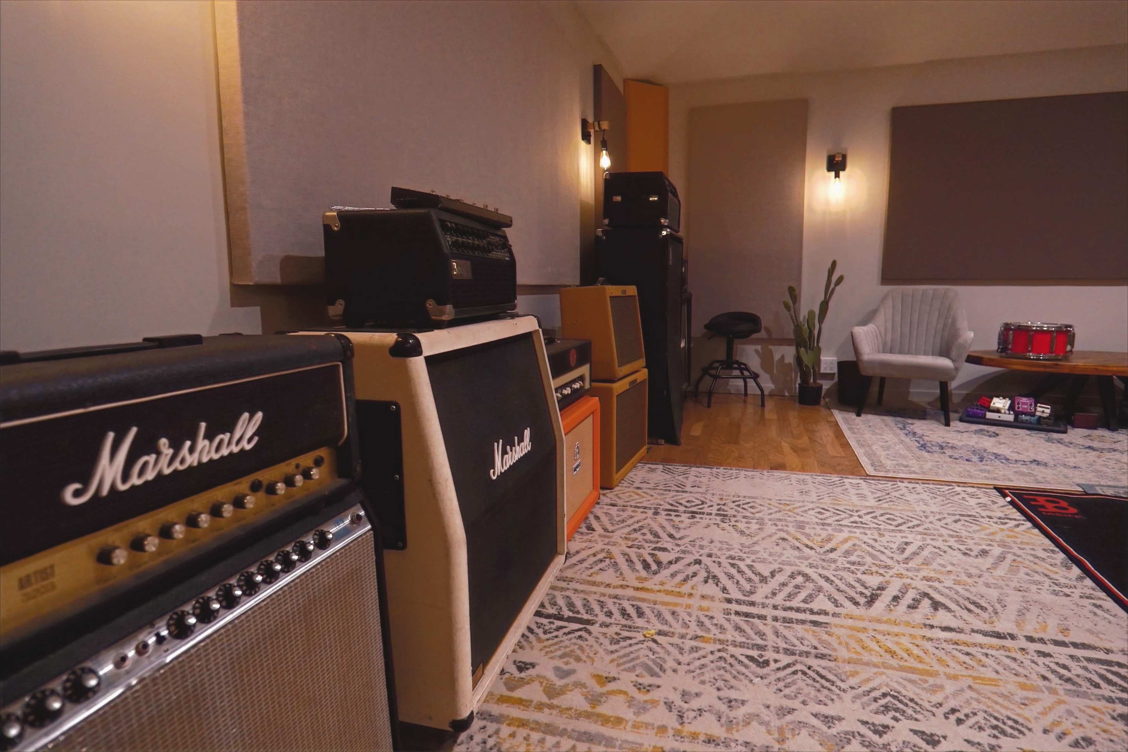 A music studio featuring several amplifiers stacked against the wall, a chair, a drum kit, and soundproofing panels.