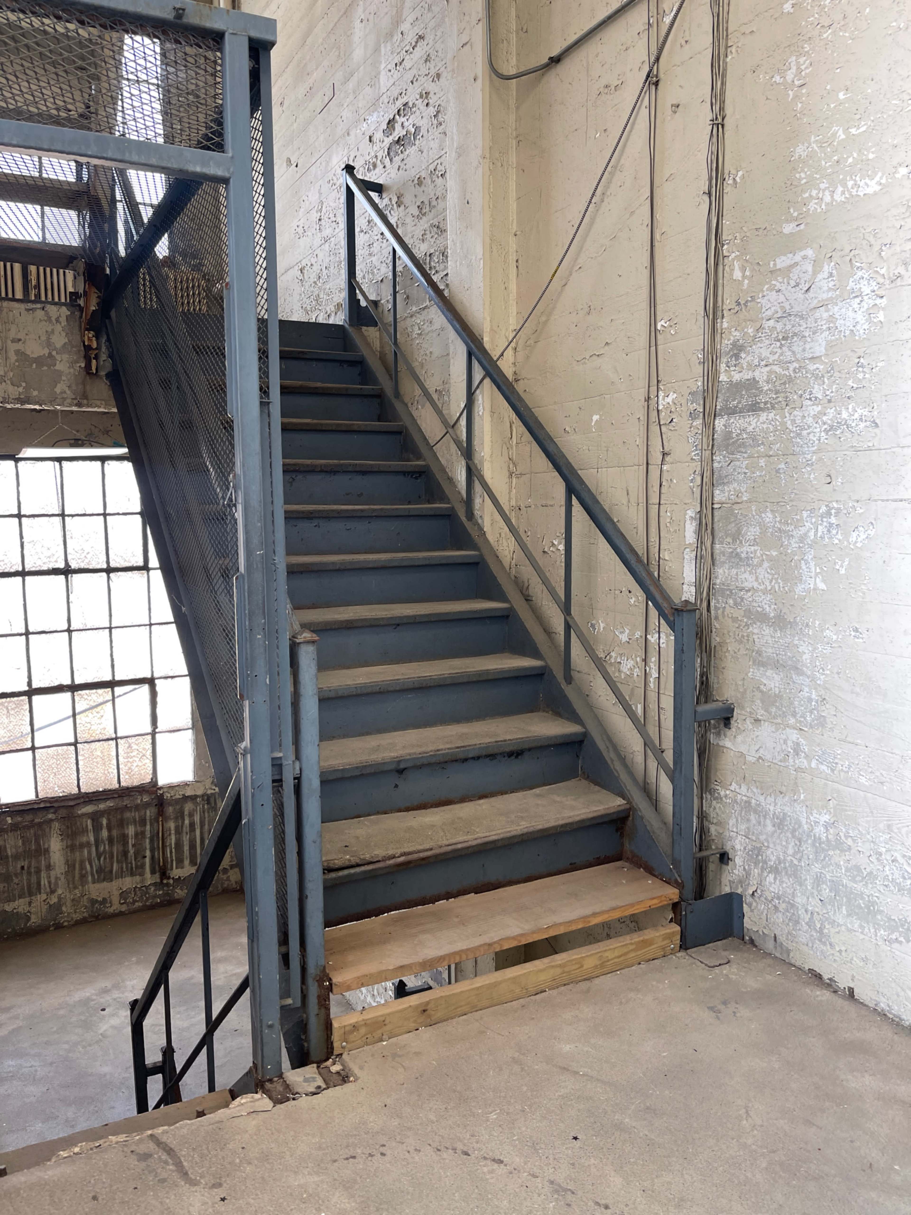 A metal staircase leads to an upper level in a corridor with bare concrete walls.