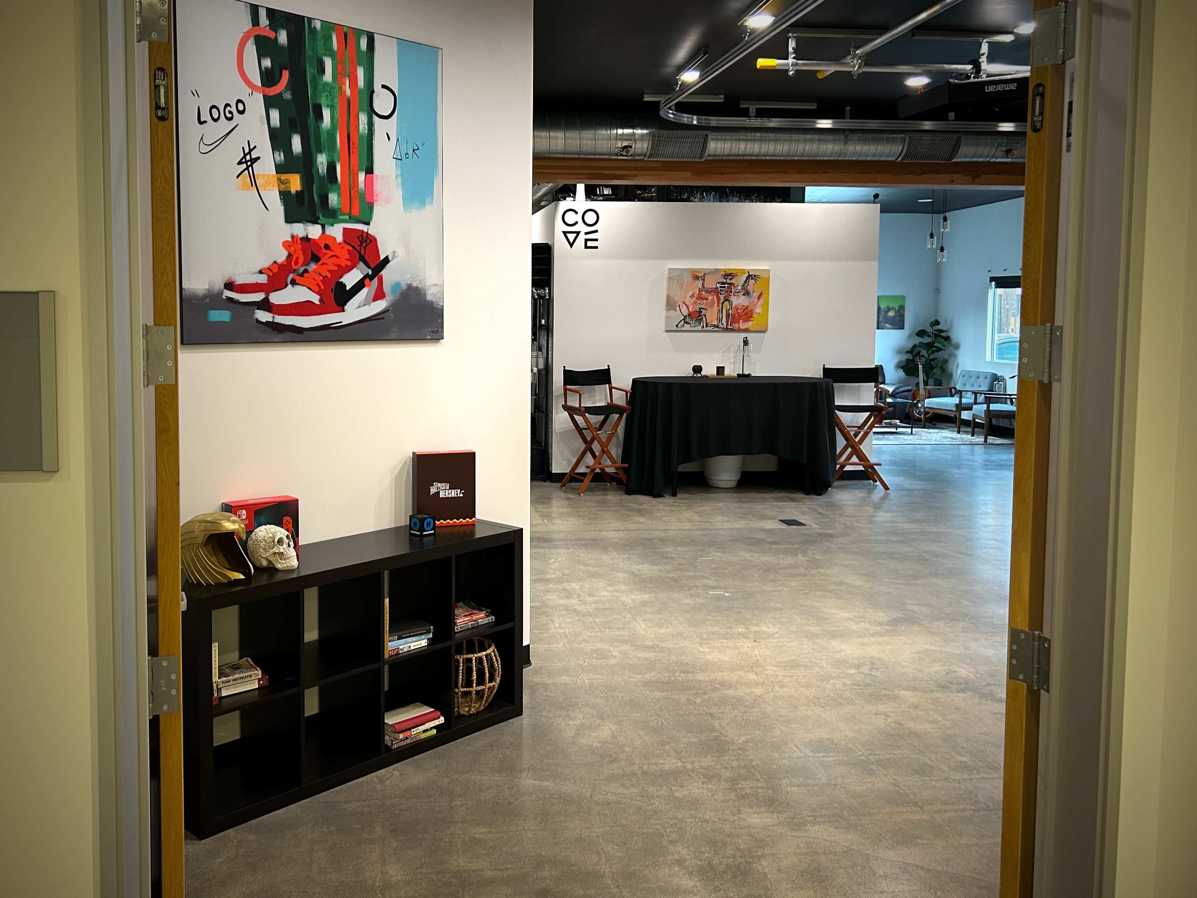 A hallway leads to a spacious room featuring a black table, chairs, and artwork on the walls.
