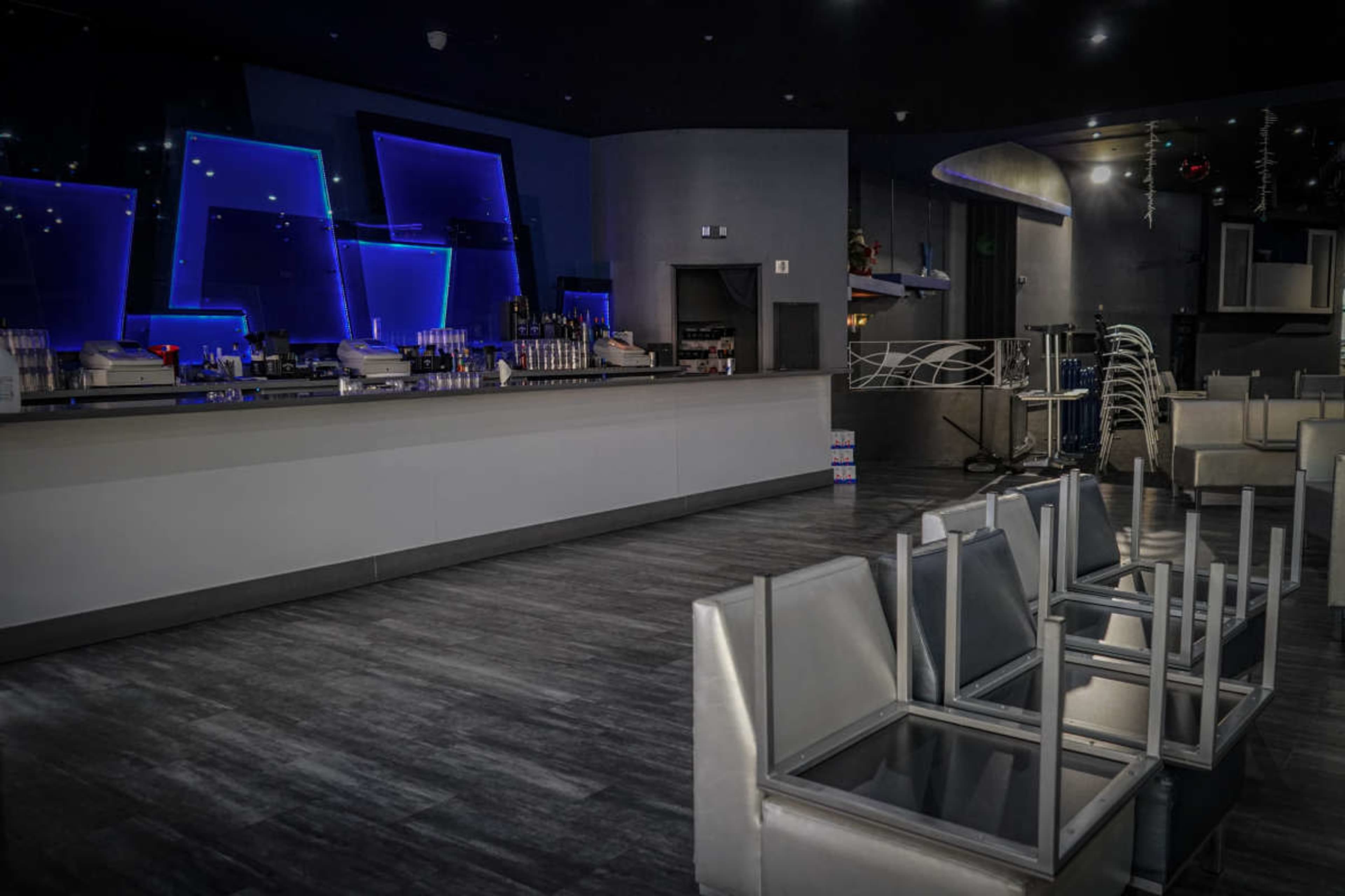 The image shows a modern bar area with blue neon lighting, empty chairs stacked on tables, and a sleek counter in a dimly lit space.