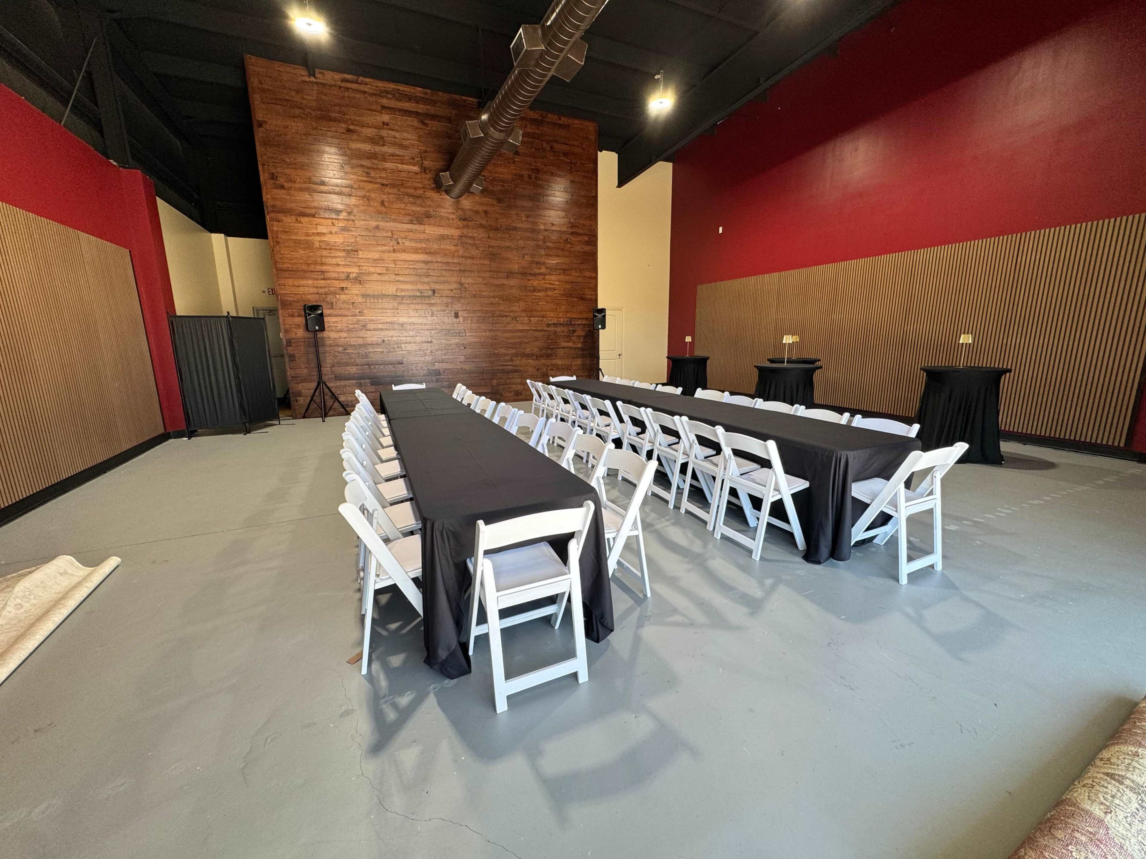 The image shows a large, open event space with rows of chairs and tables set up in a rectangular arrangement, featuring a wooden wall at one end and a red accent wall.