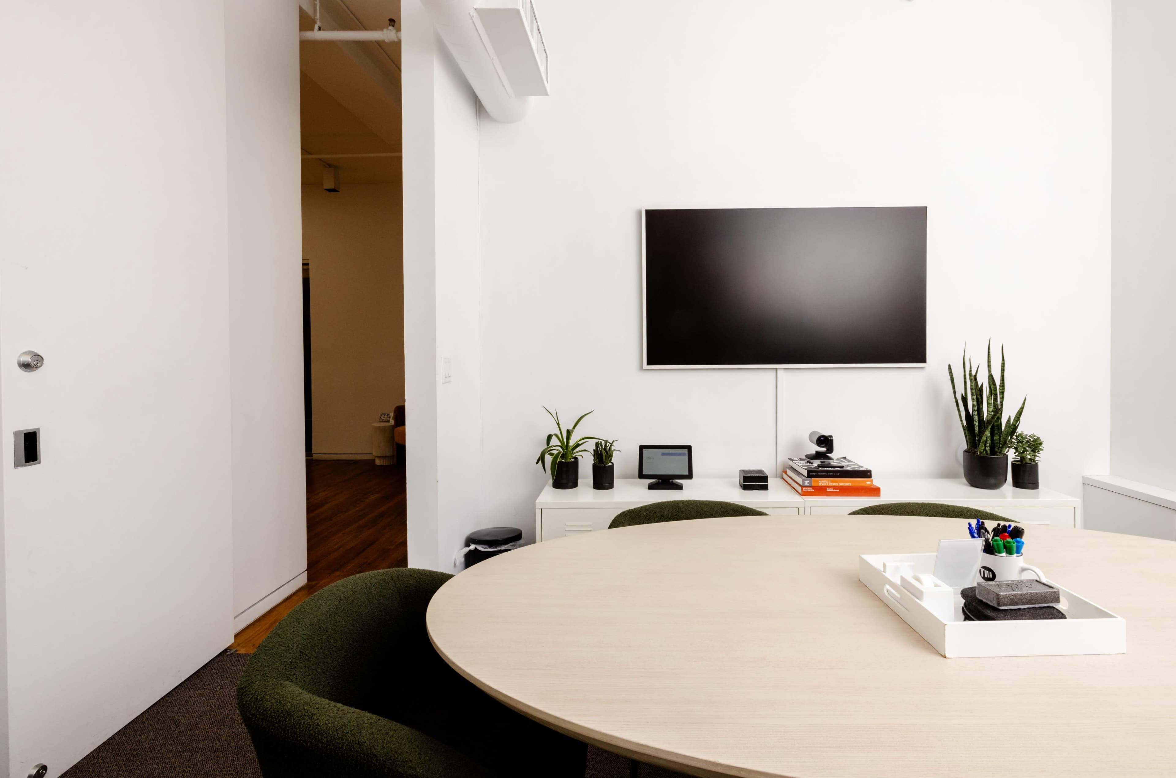 The image shows a modern meeting room with a round wooden table, a television mounted on the wall, and green plants in pots.