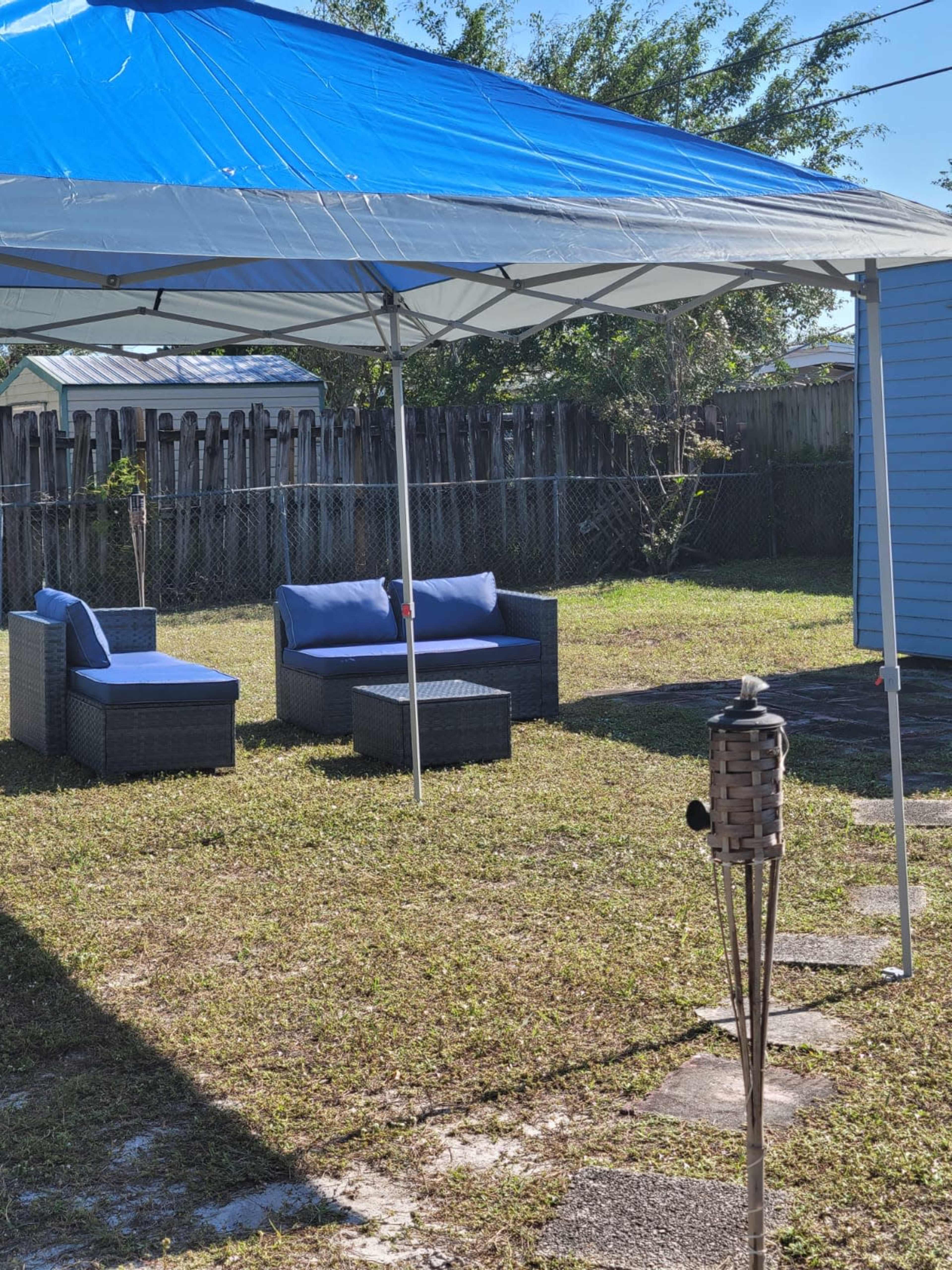 A shaded seating area with blue cushioned chairs and a small table is set up under a canopy in a grassy backyard.