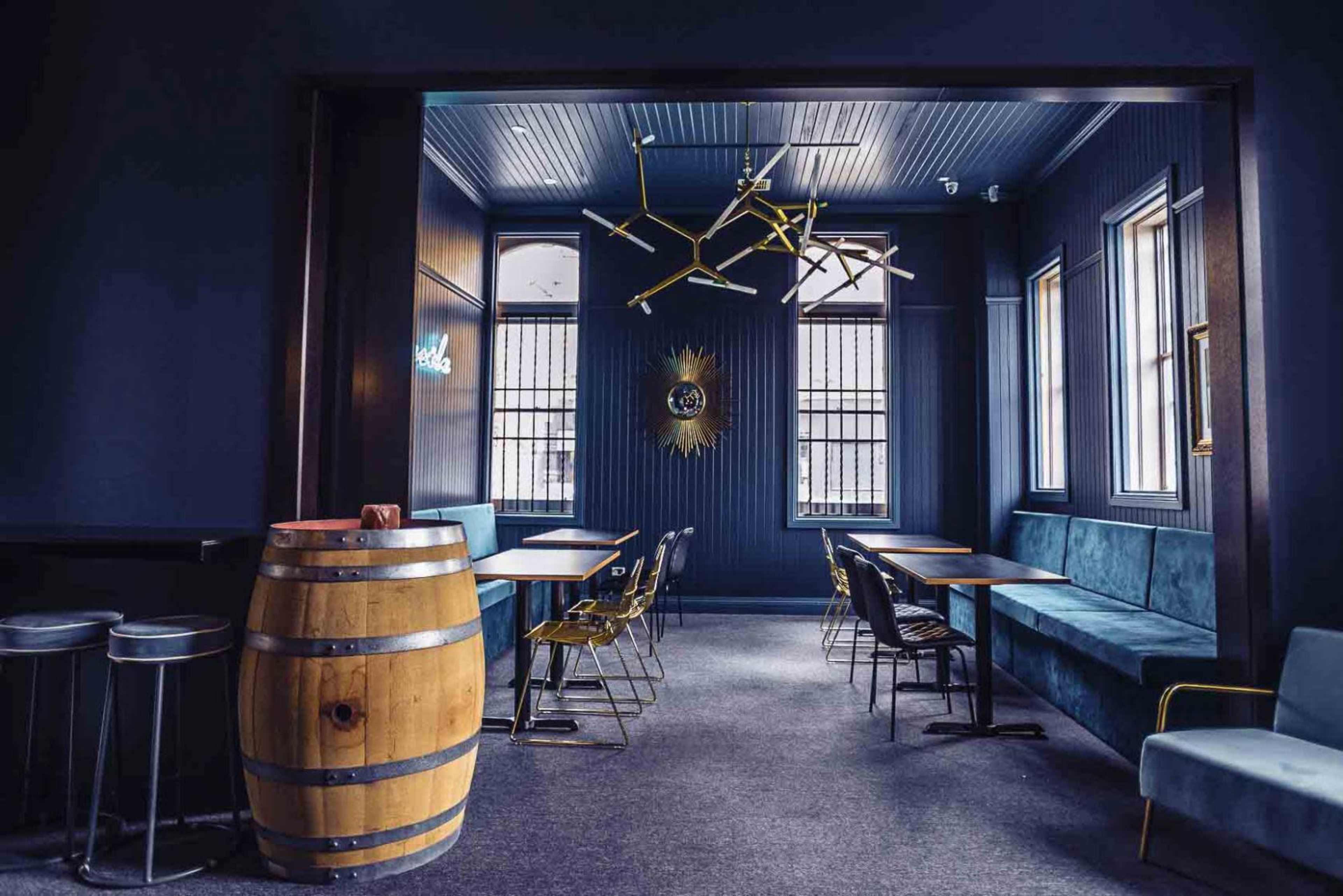 A dark-themed restaurant interior featuring a wooden barrel, metal chairs, and tables arranged in a stylish layout with large windows.