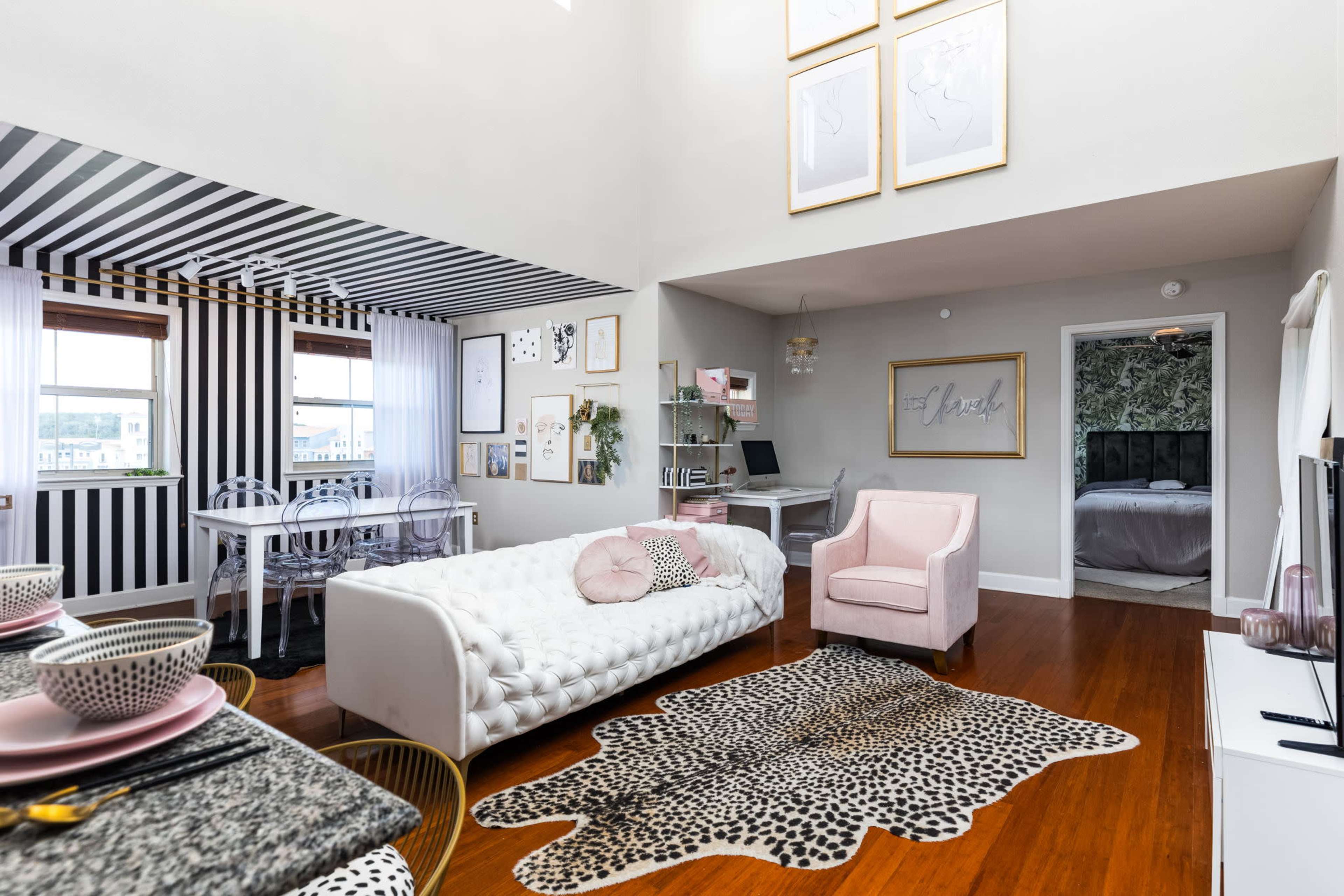 The image shows a modern living space with a striped accent wall, a white tufted sofa, a leopard print rug, and a dining area featuring a round table and chairs.