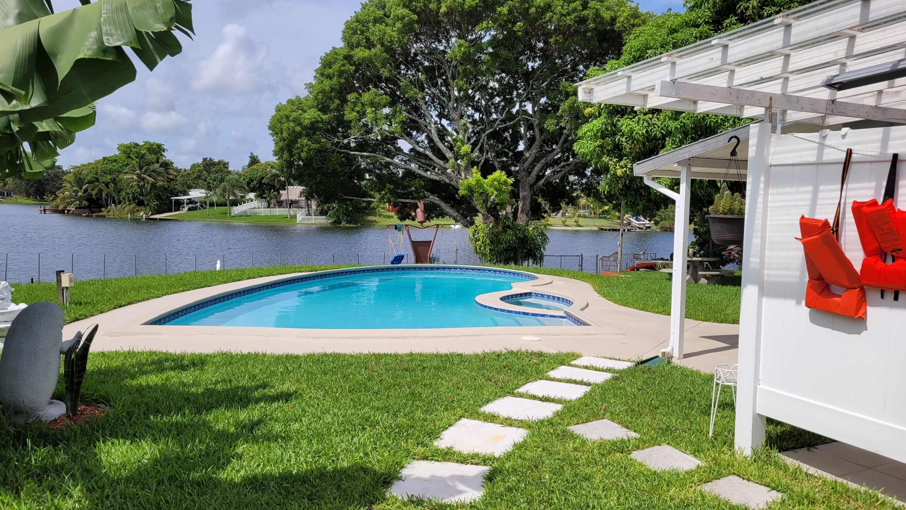 A swimming pool surrounded by grass and a white patio structure, with a large tree and a lake in the background.