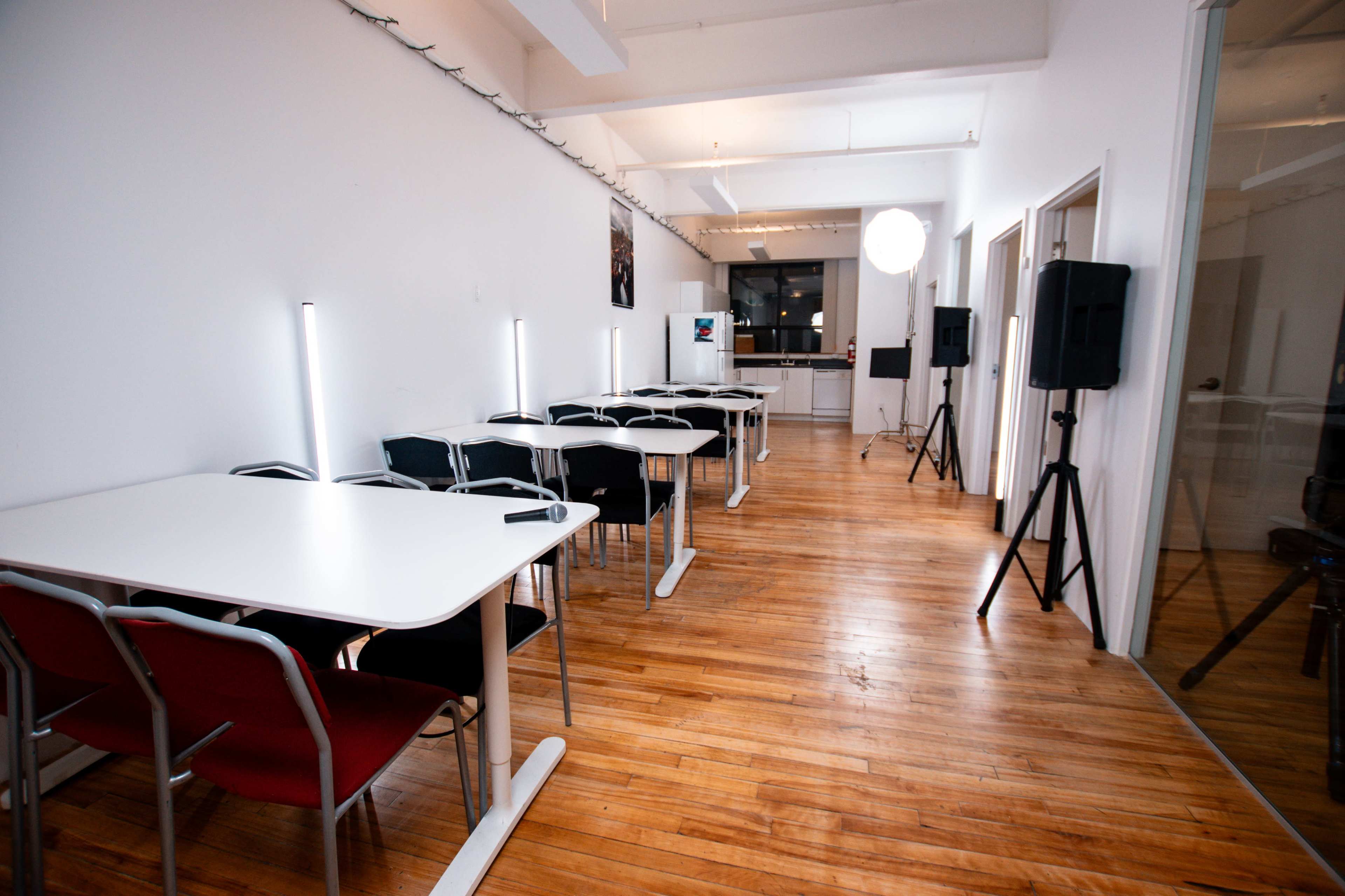 The image shows a spacious room with several tables and chairs arranged in a row, featuring wooden floors and bright white walls.