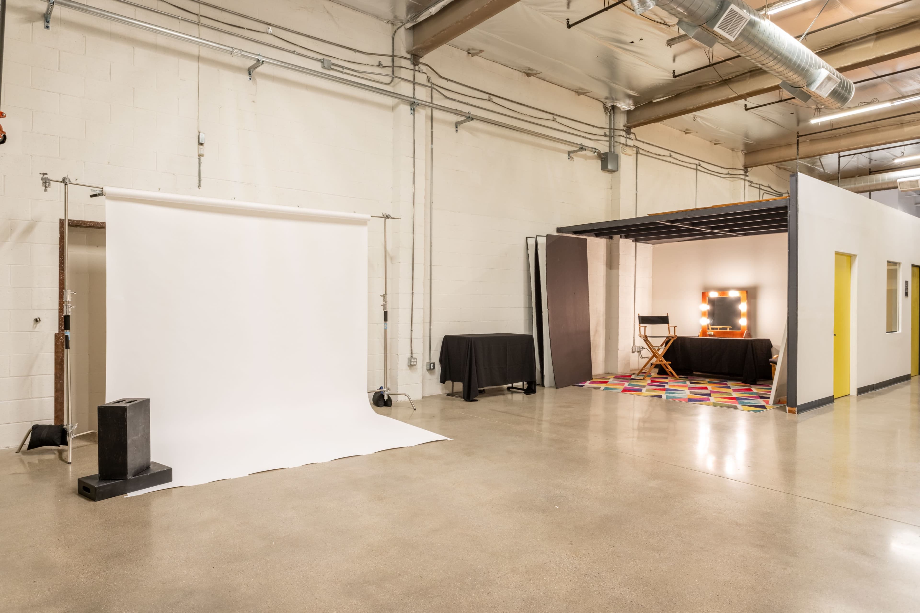 The image shows a minimalist studio space with a white backdrop on one side and a styled corner featuring a mirror with lights, a director's chair, and a colorful rug on the other.