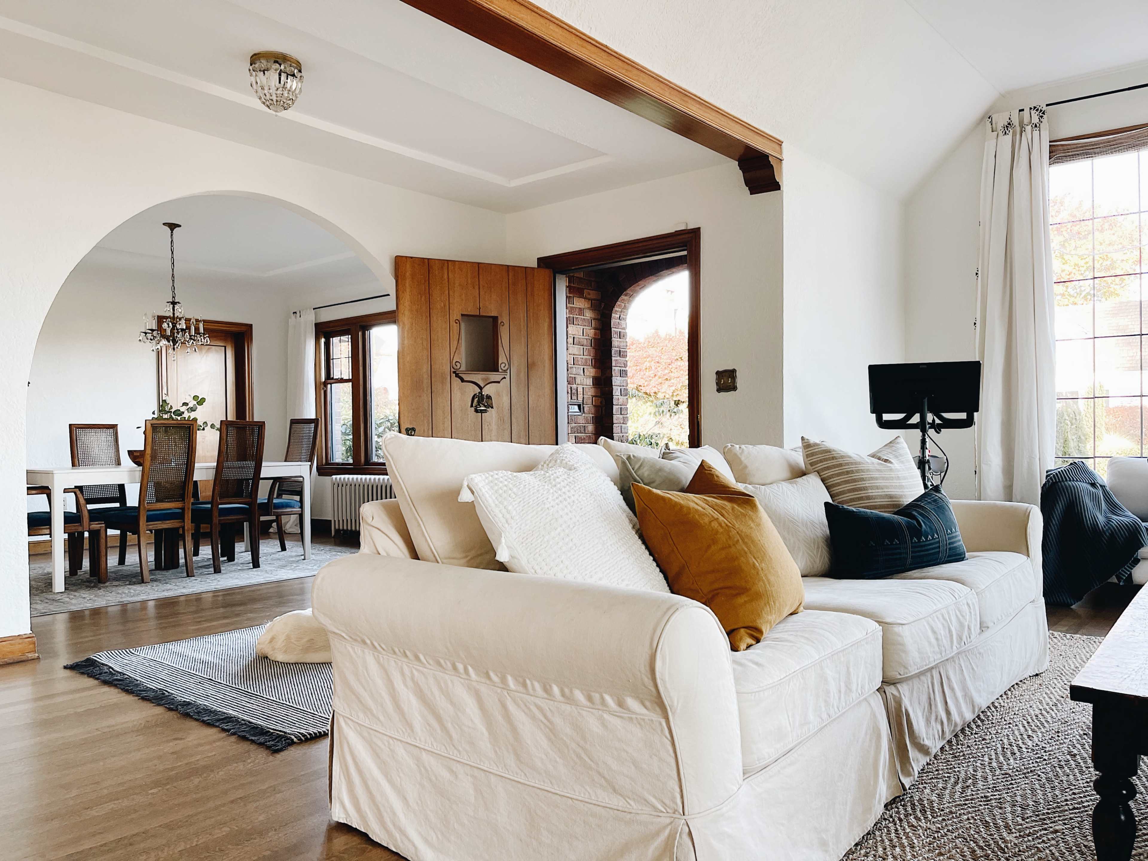 A light-filled living room features a white sofa with decorative pillows, facing an open dining area with a wooden table and chairs.