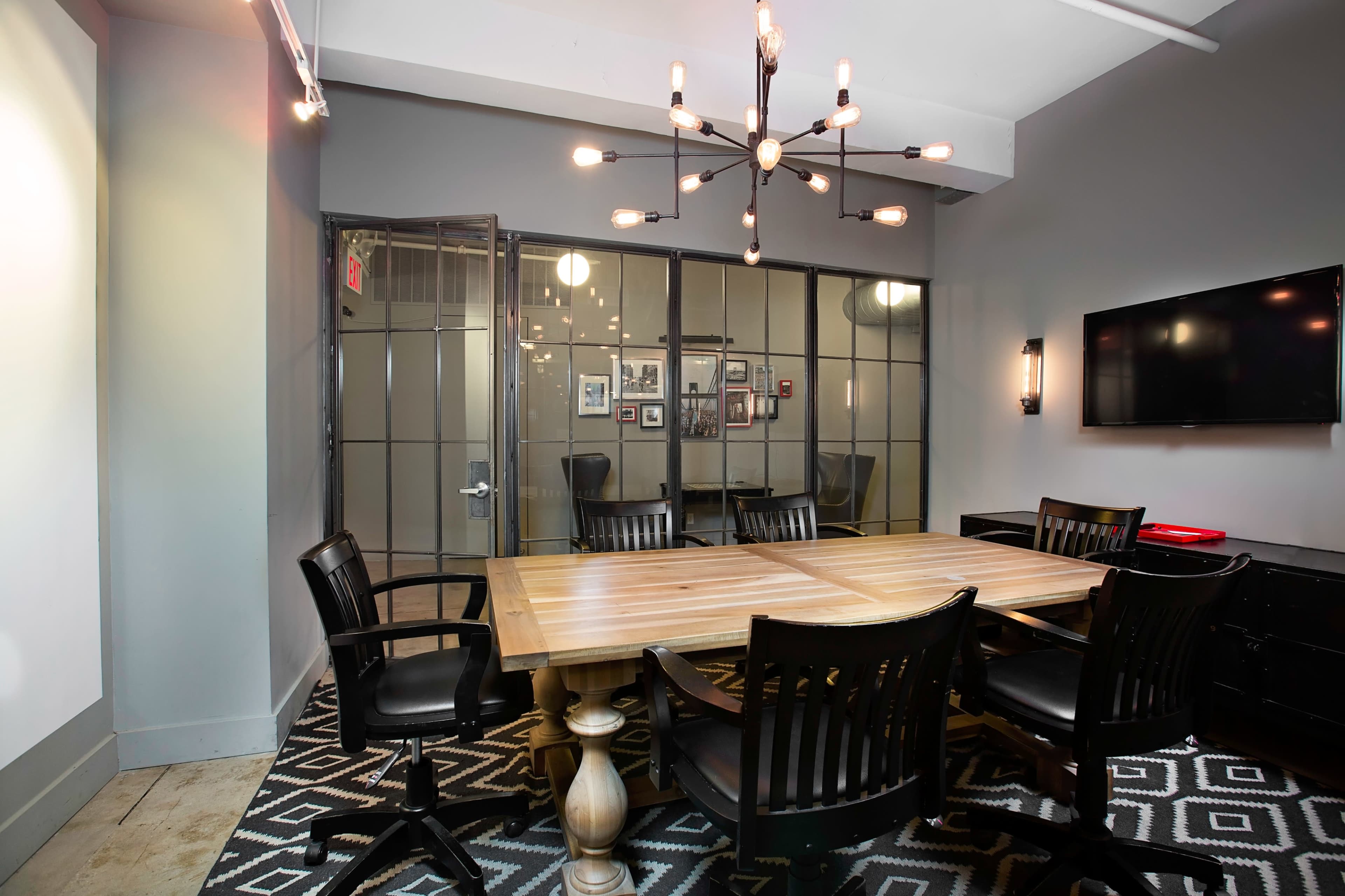 A conference room features a large wooden table surrounded by black chairs, with glass doors leading to another room and a wall-mounted television.