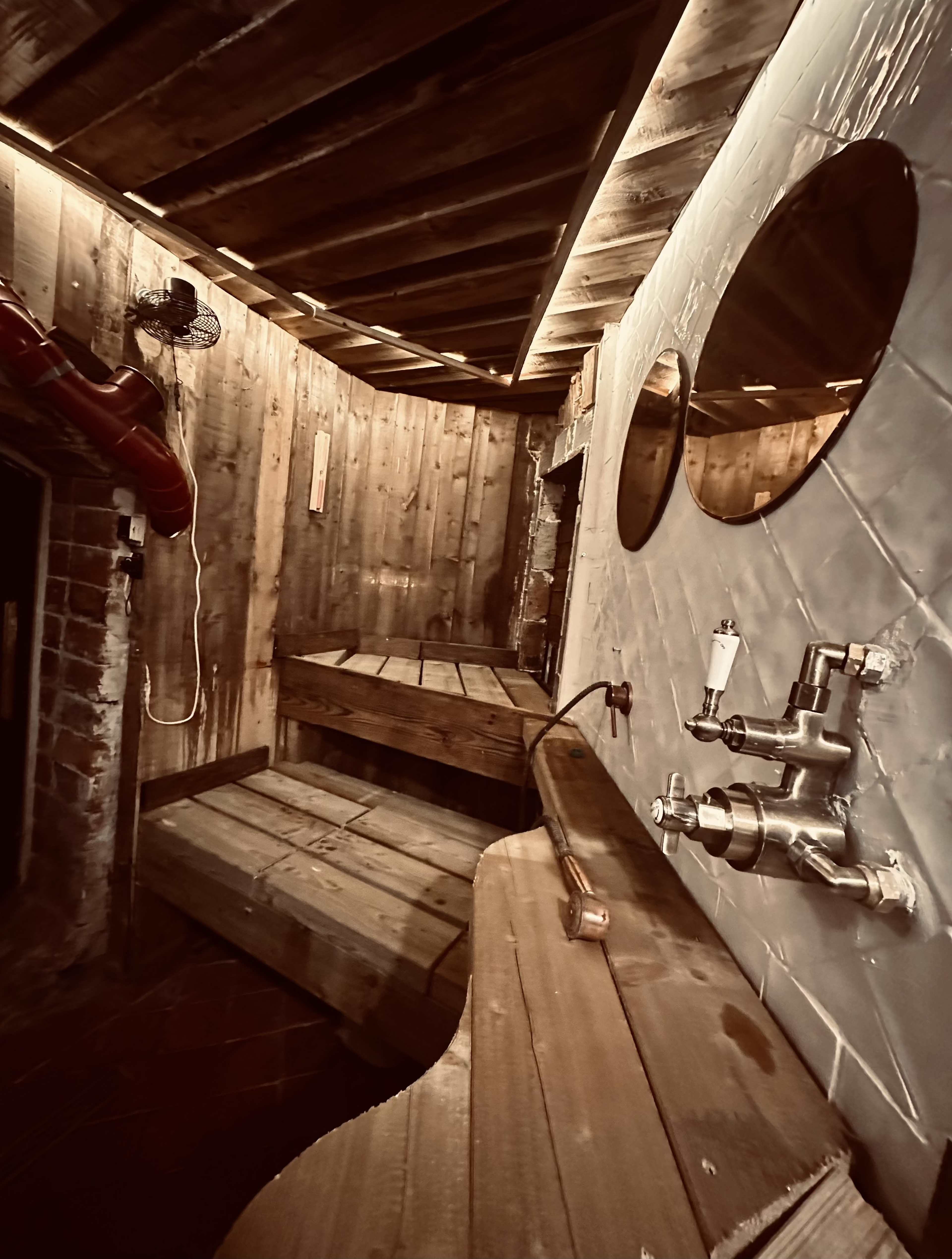 A wooden sauna interior featuring benches, two round mirrors on the wall, and various plumbing fixtures.