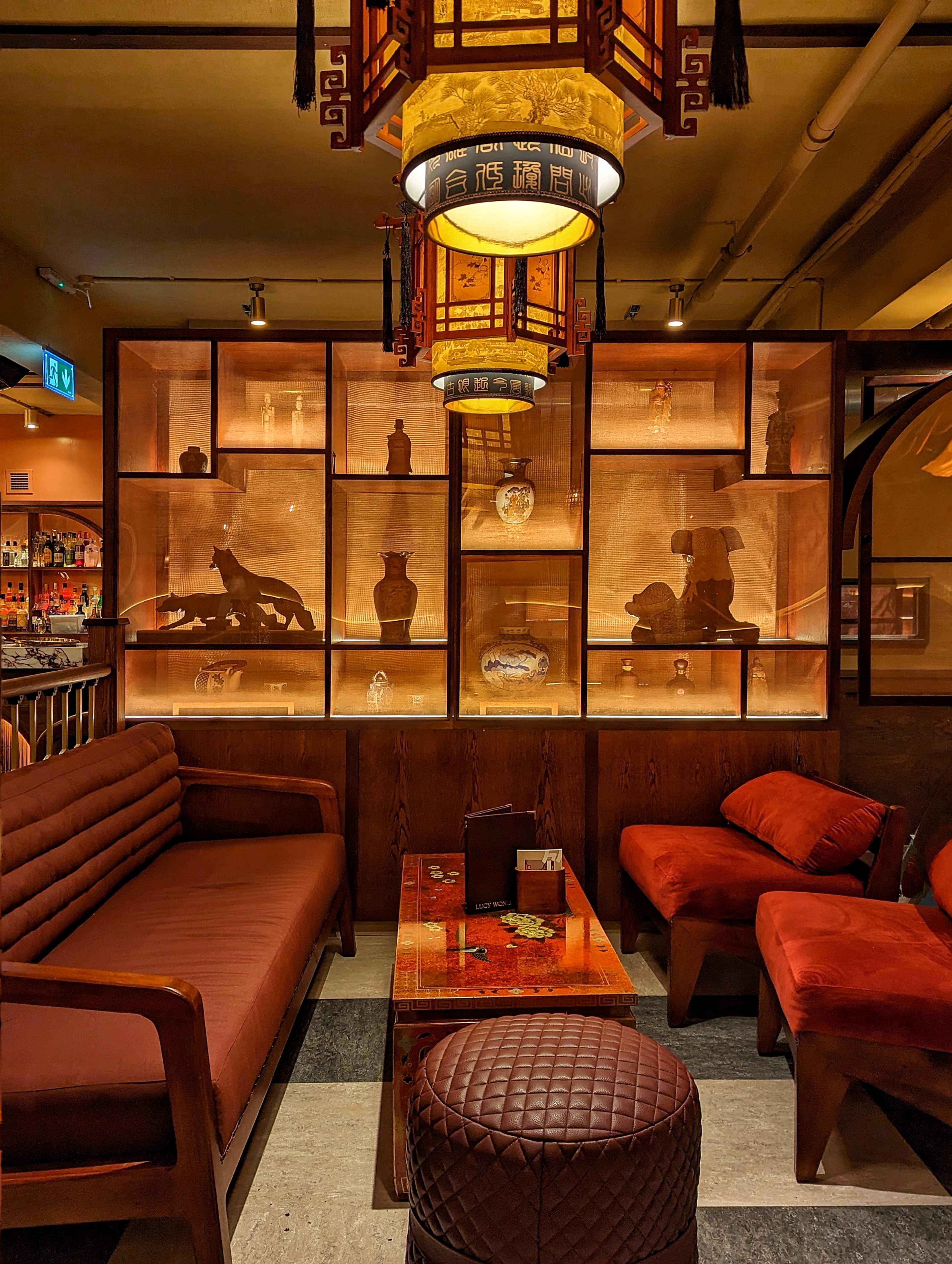 The image shows a cozy seating area in a dimly lit restaurant, featuring a wooden shelf display with illuminated artifacts and decorative items.