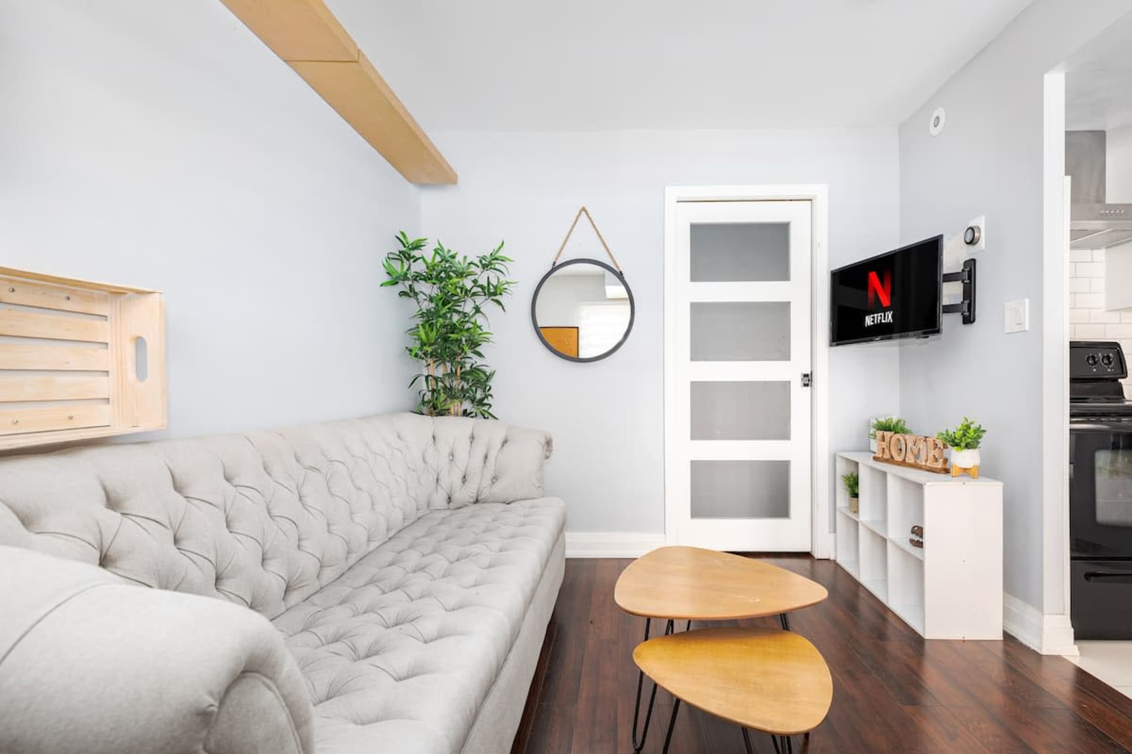 A modern living room featuring a tufted gray sofa, a round mirror, a small wooden coffee table, and a wall-mounted TV displaying the Netflix logo.