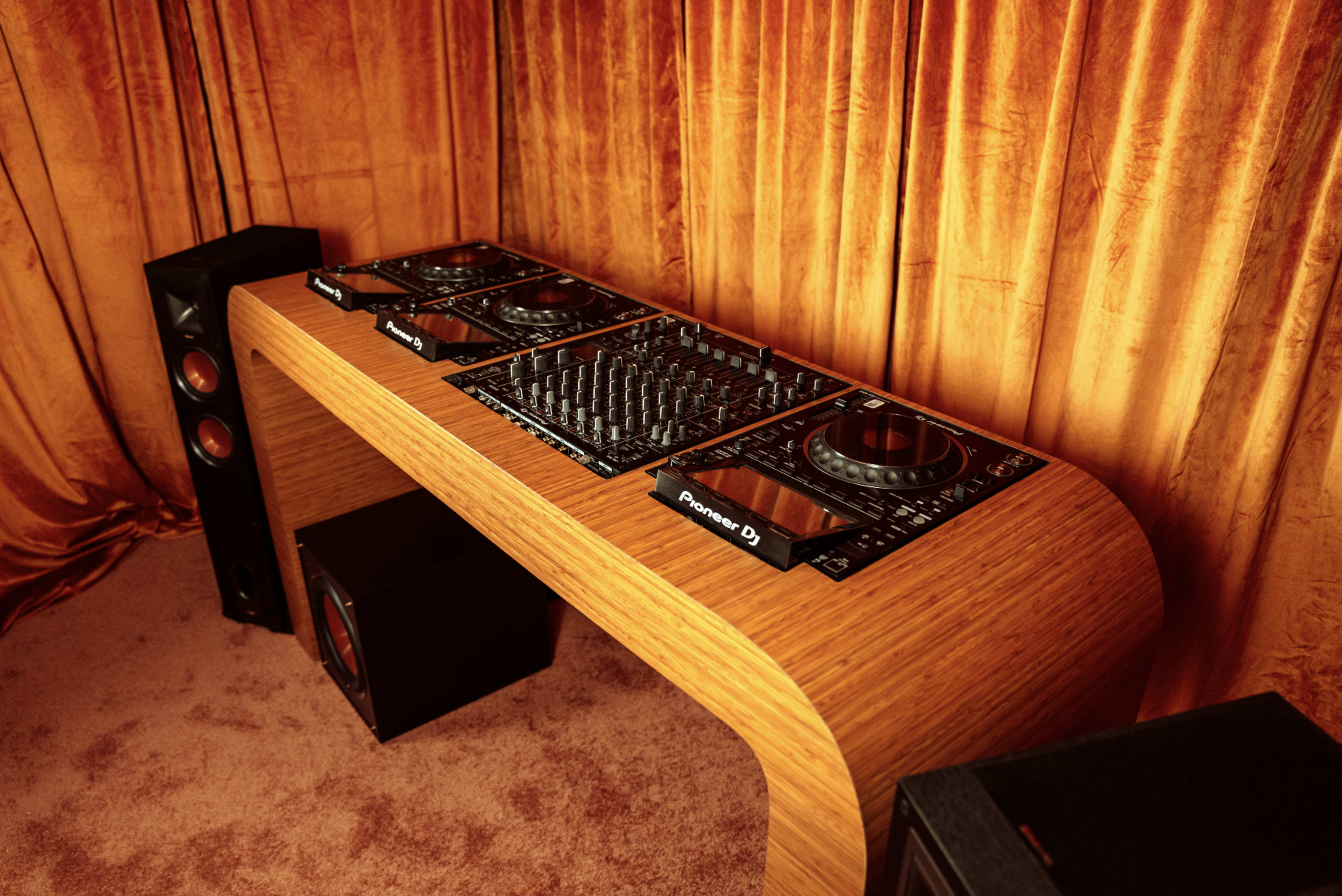 The image shows a DJ setup featuring a wooden console with turntables and mixers, flanked by tall speakers, all set against a backdrop of orange curtains.