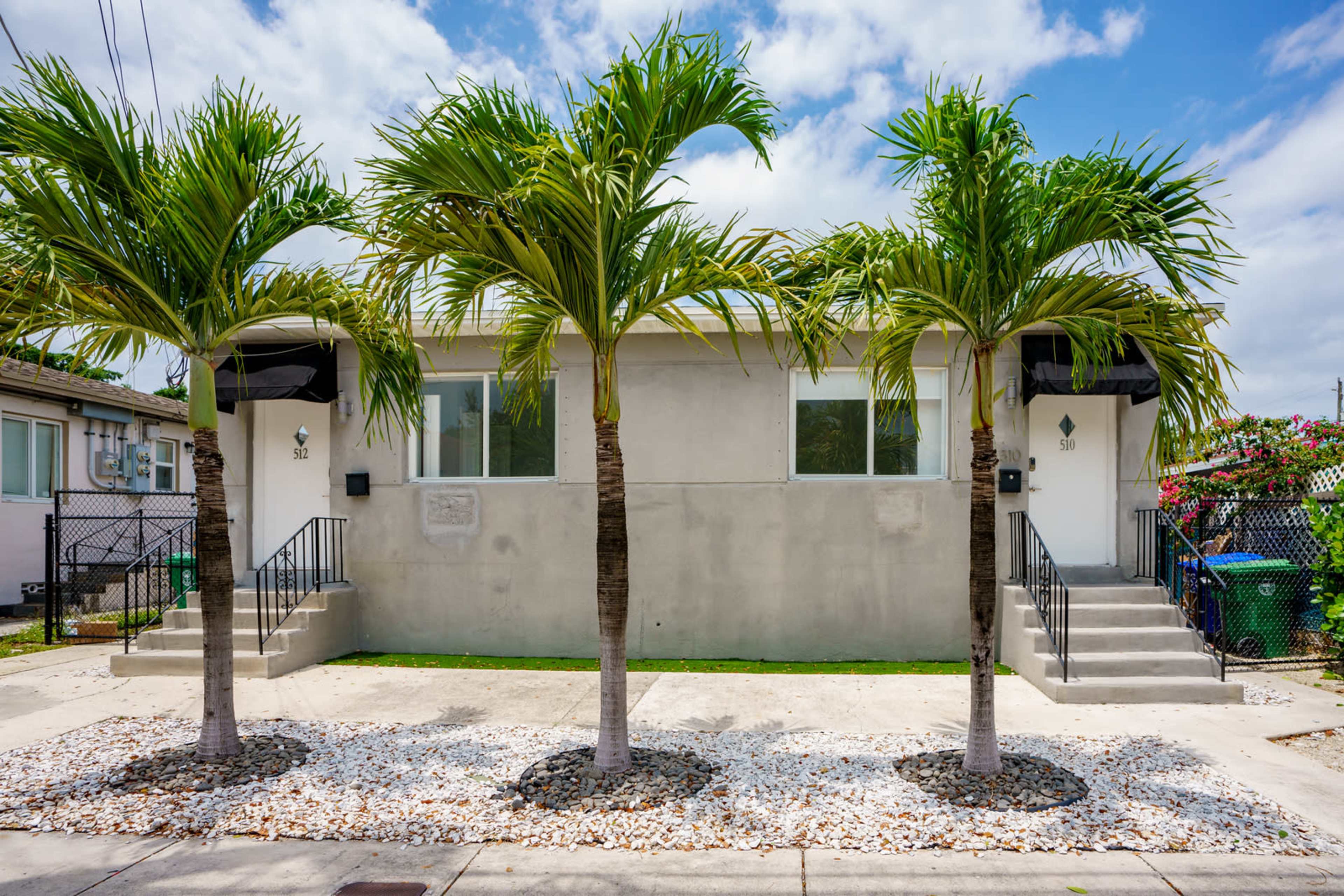 A duplex with two entrances is flanked by three palm trees and features a gravel landscaping area in front.