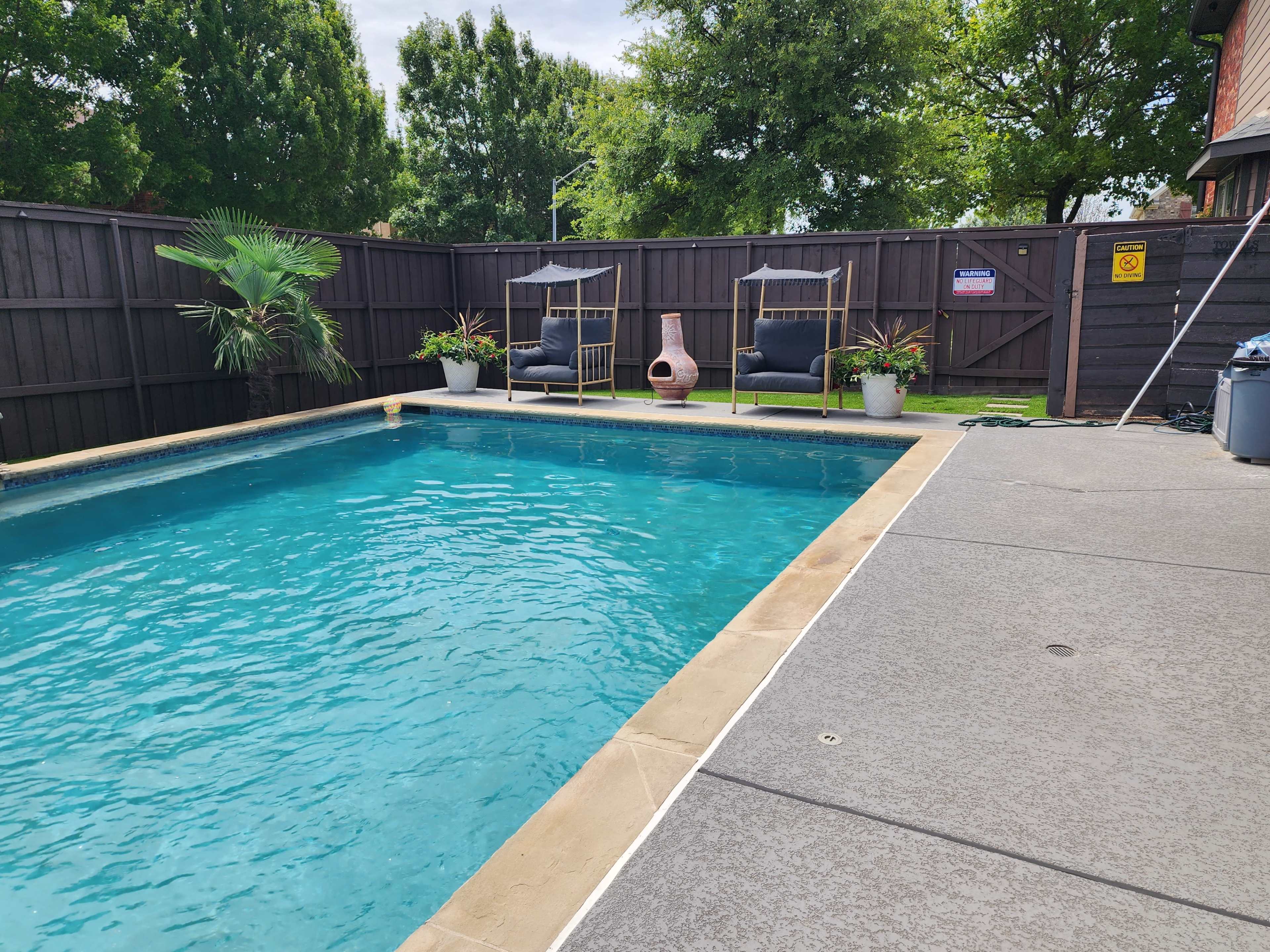 The image depicts a swimming pool surrounded by a concrete deck, with two lounge chairs and potted plants positioned nearby.