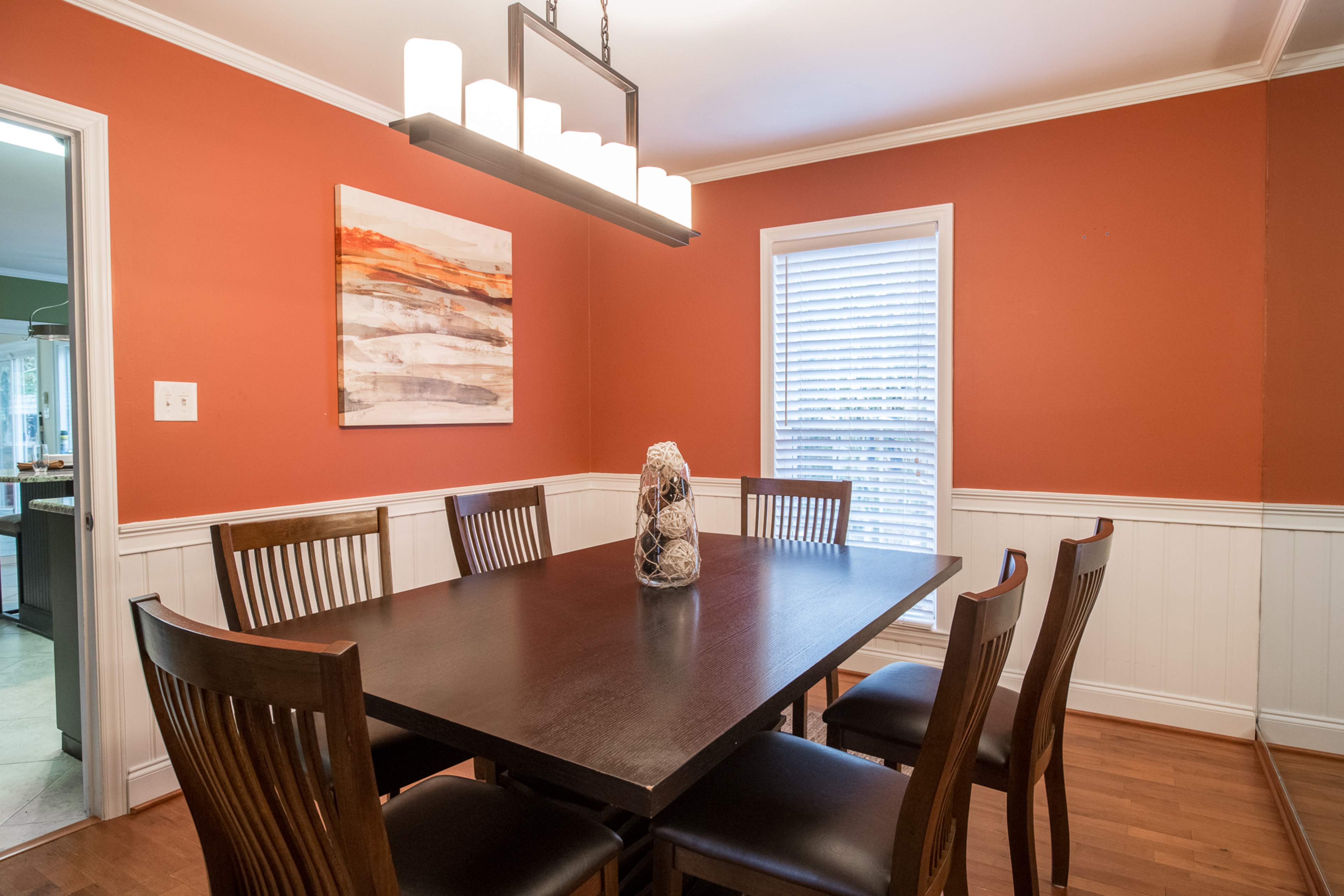 A dining room features a dark wooden table surrounded by six chairs, a pendant light overhead, and an orange accent wall with a framed artwork.