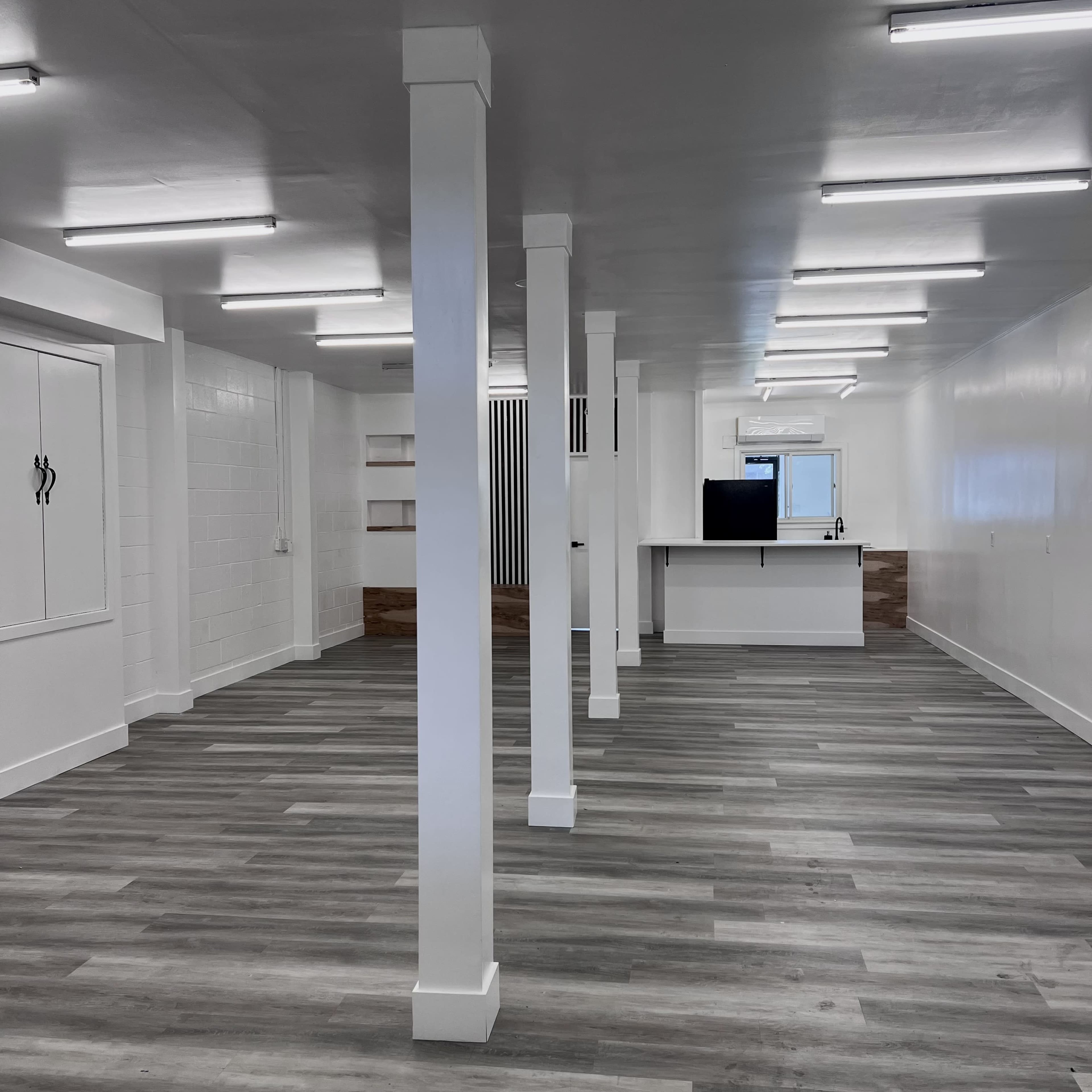 An empty room with gray wooden flooring, white walls, and several white supporting columns, featuring a kitchen area in the back.