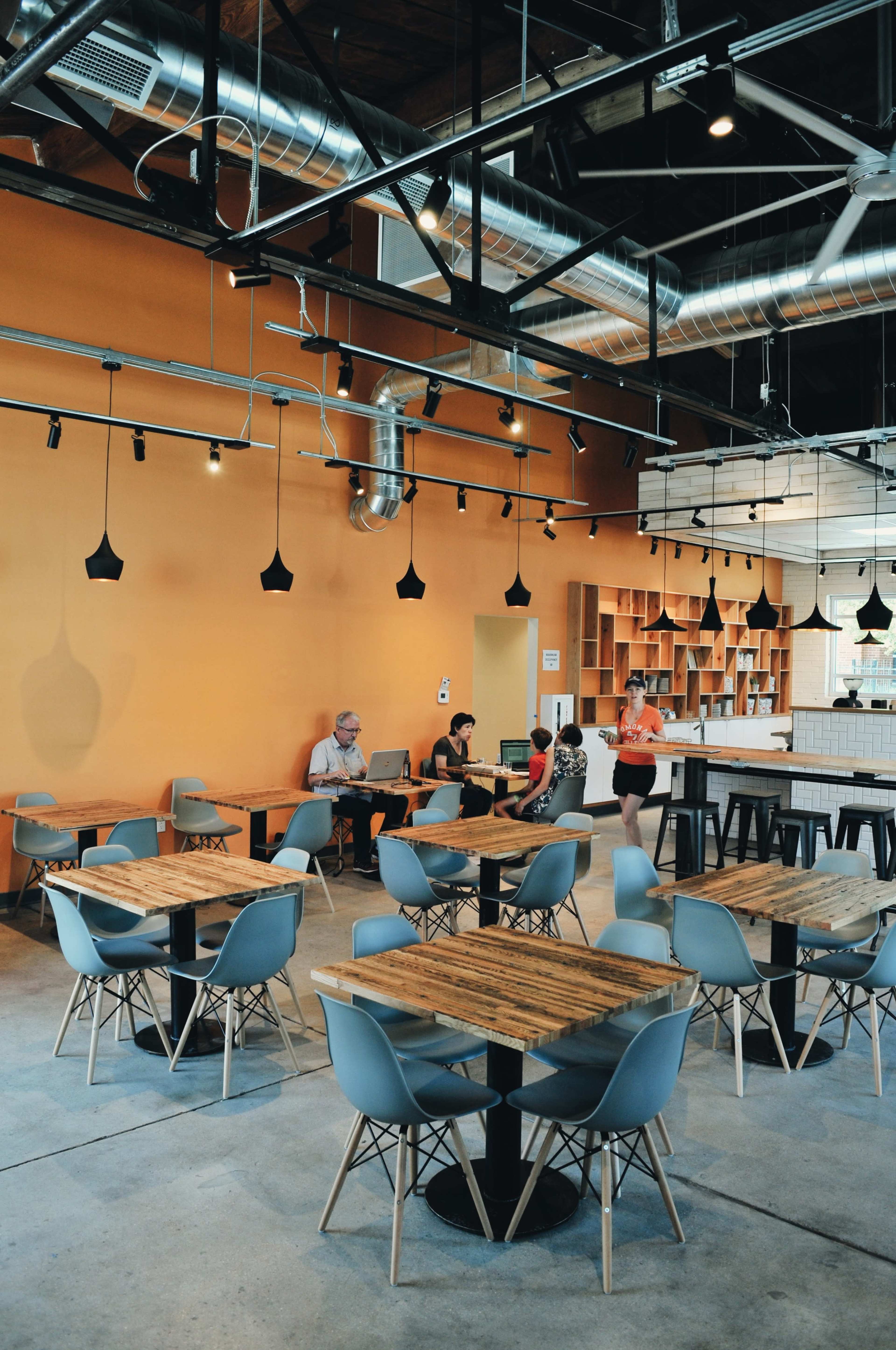 A modern cafe features wooden tables and gray chairs, with a group of people seated at a central table and an orange wall in the background.