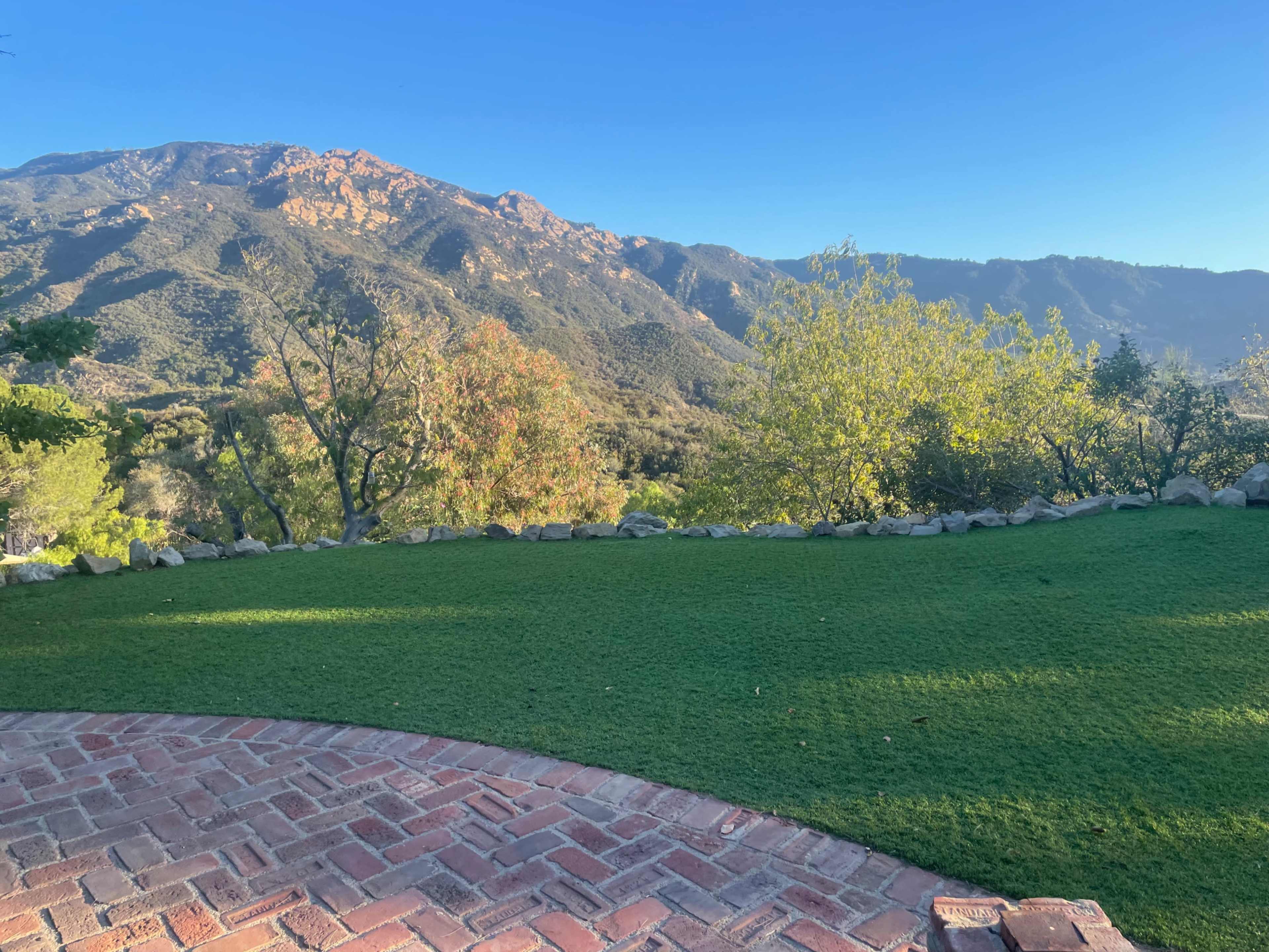 A manicured lawn sits in the foreground, framed by a stone wall with mountains and trees extending into the background.