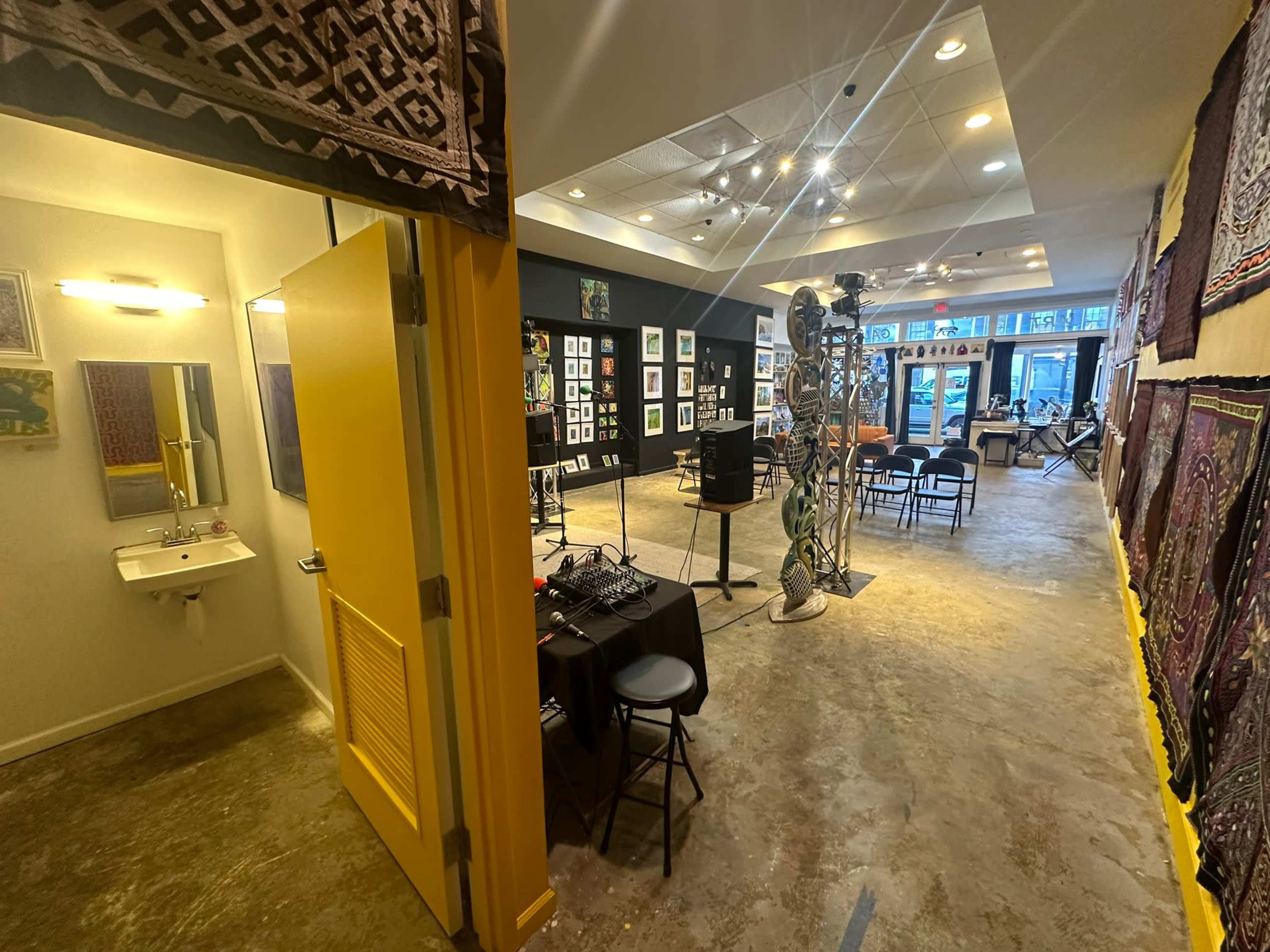 An art gallery interior featuring artwork on the walls, a yellow door leading to a restroom, and seating arranged for visitors.
